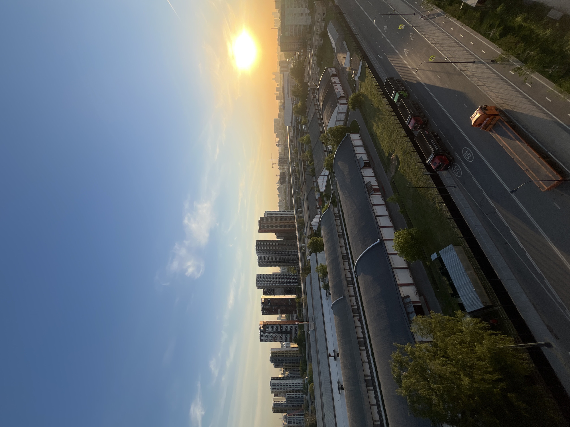An elevated perspective shows a city view at dusk, likely in Moscow, Russia, given the architecture. The horizon is bathed in golden light from the setting sun, with a clear blue sky. Below, a major highway runs alongside industrial buildings with curved roofs. A long, flatbed truck carrying heavy machinery travels along the highway. Residential buildings can be seen, too.
