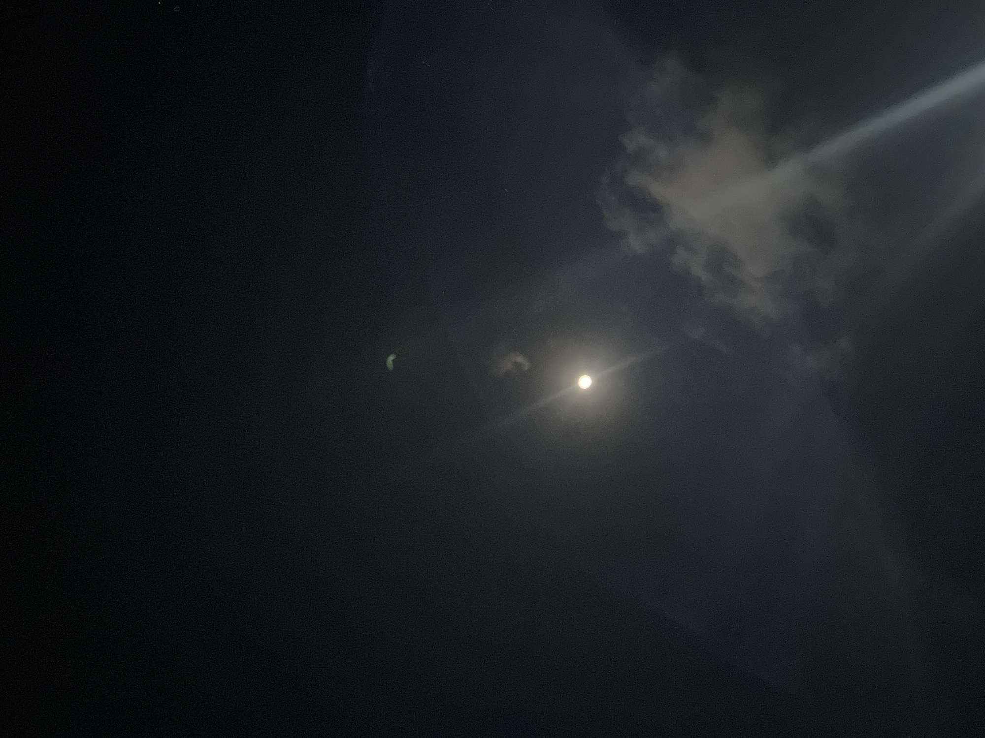 A nighttime image shows the moon in the sky, partially obscured by clouds. The moon appears as a bright, circular light source. There are also visible clouds illuminated by the moonlight, creating streaks of light. The overall scene is dark, suggesting it is a clear night with some cloud cover. The image was taken in Denpasar, Indonesia.