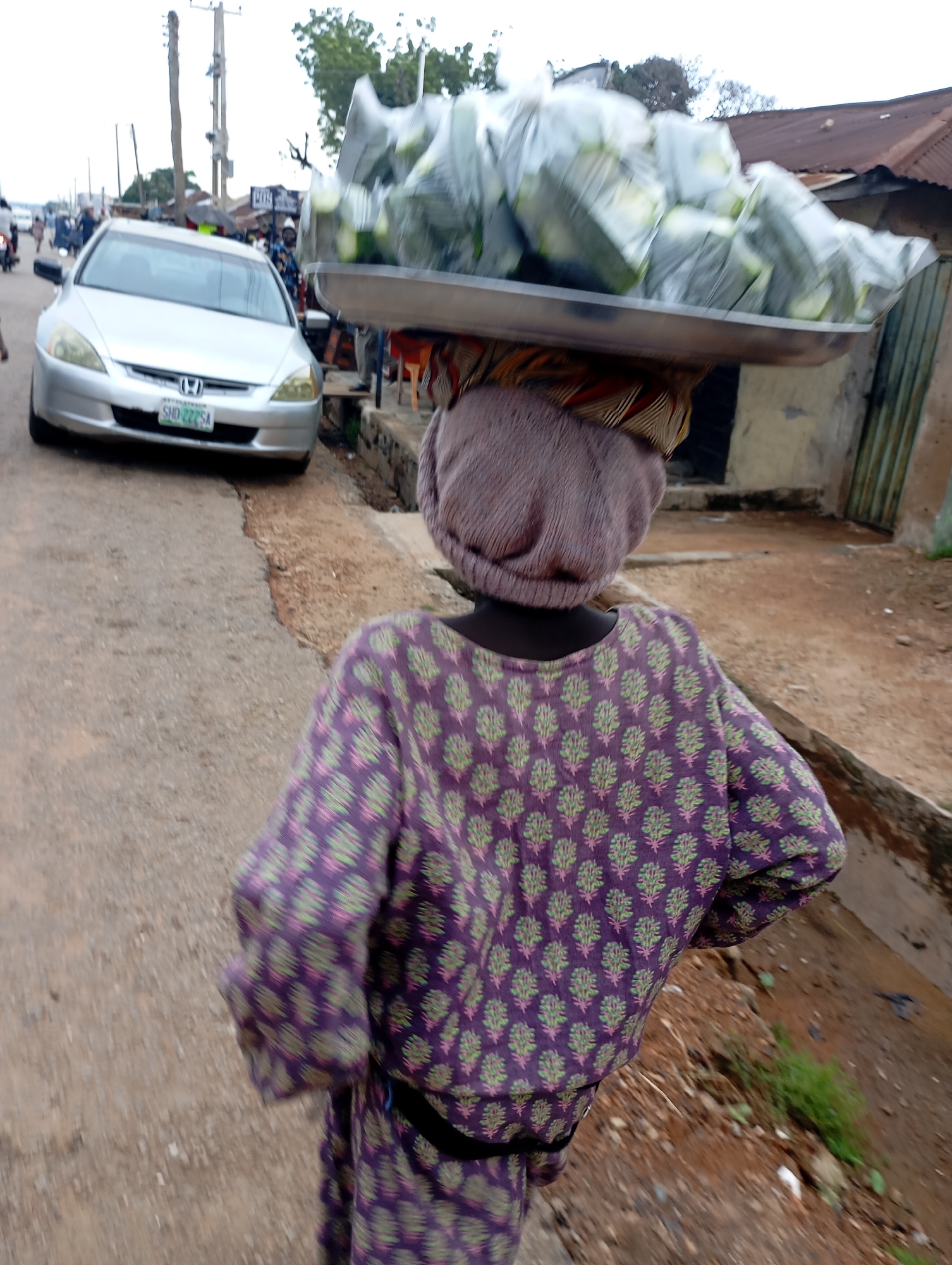 A blurred, medium shot from behind shows a woman walking on a dirt road in Shendam, Nigeria. She is wearing a purple and green patterned dress and a headscarf. On her head, she carries a large metal tray filled with plastic-wrapped bundles, likely produce. To her left, a silver sedan is parked on the roadside. In the background, buildings with corrugated metal roofs and utility poles are visible. The overall impression is a busy street scene in a Nigerian town.