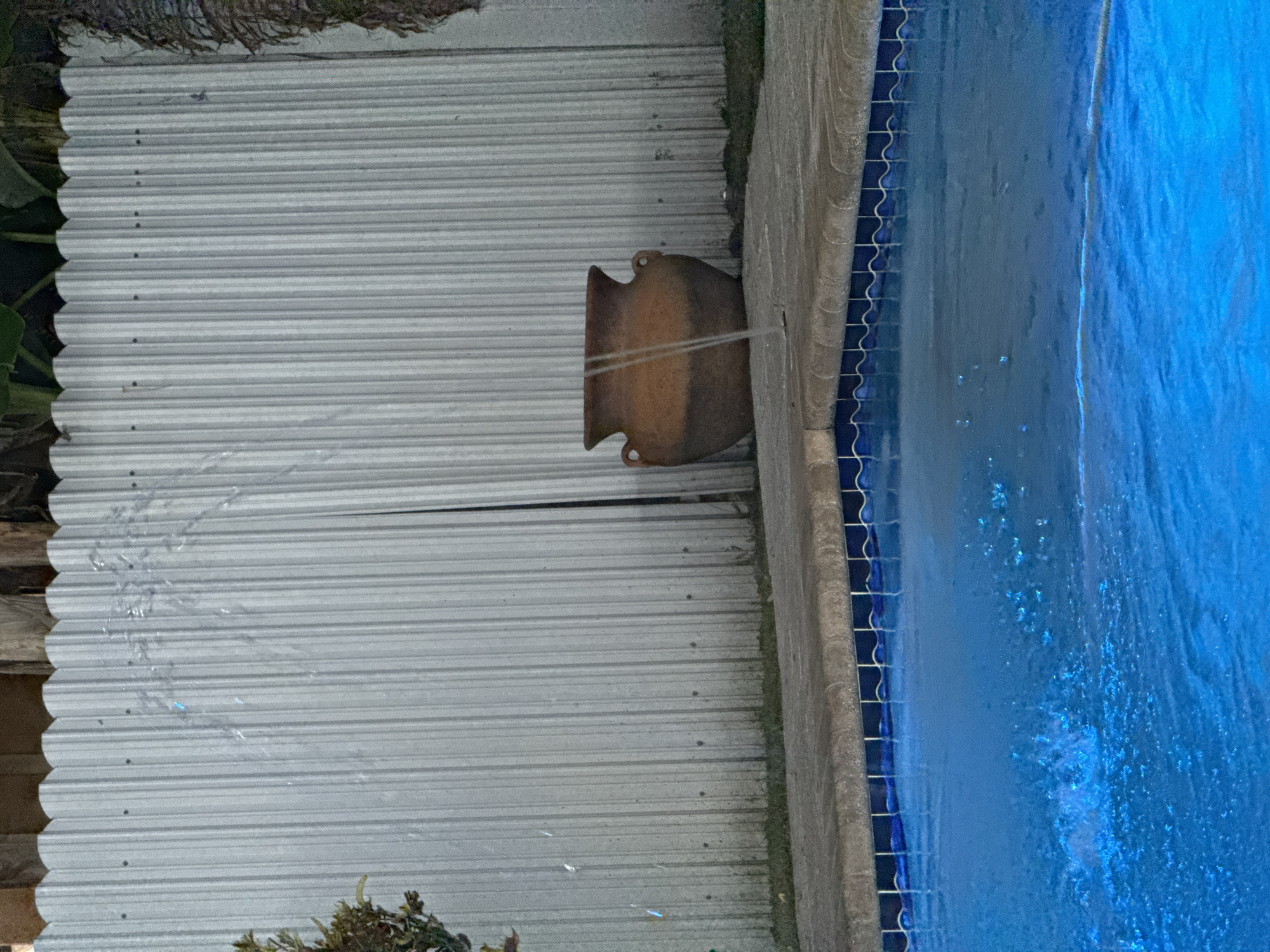 The image captures a scene near a swimming pool, likely in Miami, United States of America, given the typical South Florida style of pool construction and landscaping. A large, brown, decorative pot or urn is positioned on the pool's edge, seemingly used as a water feature, as water streams are visible exiting the pot and flowing into the pool. The pool itself has a bright blue interior and a dark blue tiled border. Adjacent to the pot and the pool is a corrugated metal wall. Some greenery is visible in the lower left and upper left corners of the frame. The entire image is rotated 90 degrees clockwise.
