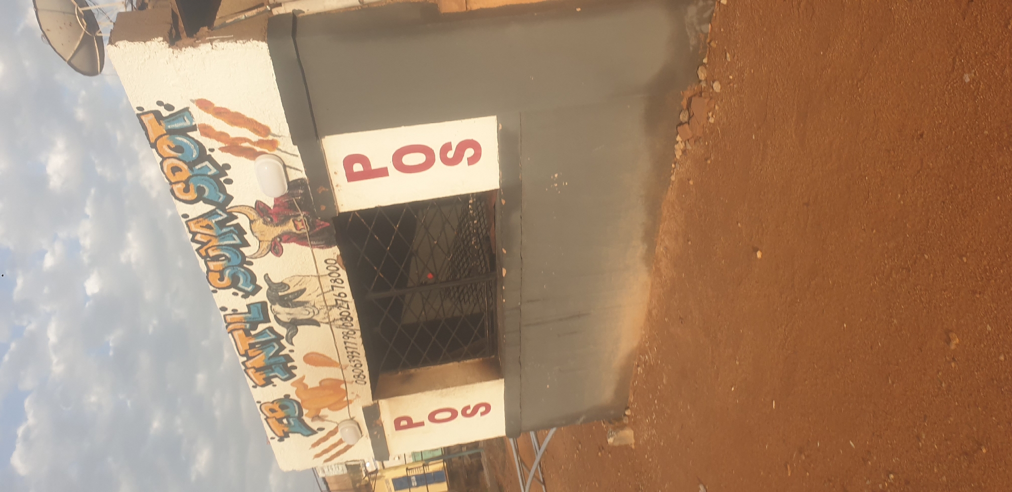 The image shows a roadside food stall with "FB INTL SUYA SPOT" painted in graffiti on its facade. There are drawings of animals, including a chicken, a goat, and a bull, along with depictions of suya (grilled meat skewers). The stall has a window with a metal grill, and "POS" is written vertically in red on a white background on the left and right sides of the stall. A satellite dish is visible on the roof. The ground in front of the stall is dirt, and there are some scattered rocks. The stall appears to be located in an urban environment, possibly in the city of Jos, Nigeria, given the mention of "SUYA SPOT" which is a popular Nigerian street food. The sky is cloudy.