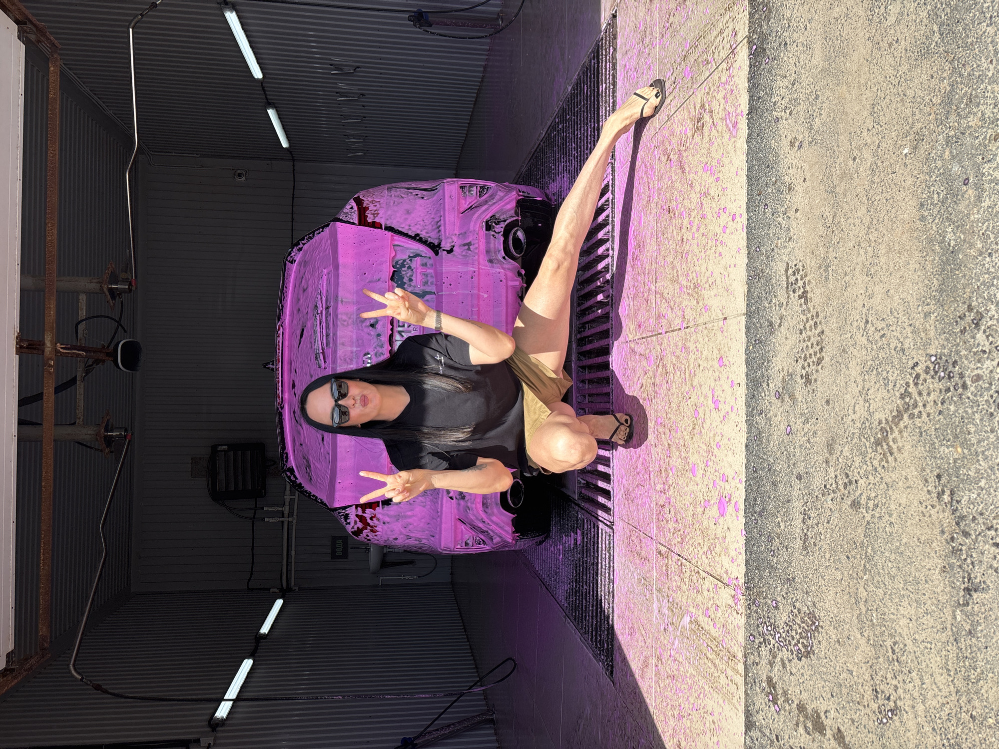A woman is posing at a car wash, possibly in Chaikovsky, Russia. She is wearing a black t-shirt, khaki shorts, and sunglasses. She is making a peace sign with both hands. Her long hair is black. The car behind her is covered in pink foam. The car wash has a corrugated metal structure and fluorescent lighting. The floor is concrete with a grate. There is pink overspray on the ground.