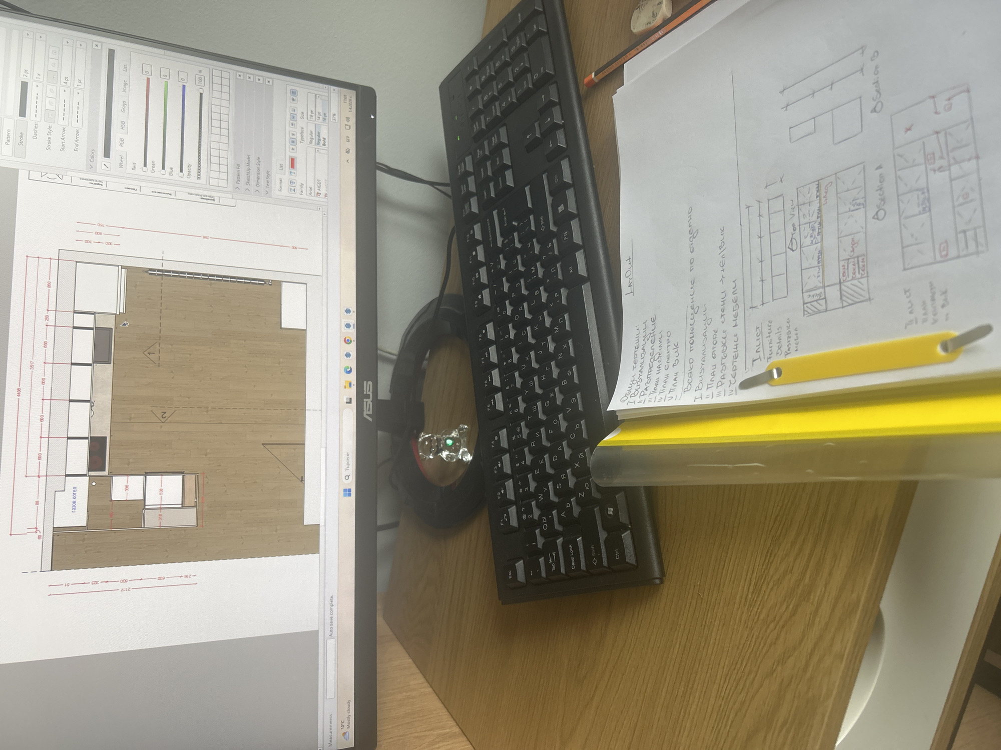 The image shows a desk with a computer monitor, keyboard, and notes. The monitor displays what appears to be architectural design software with a floor plan layout in the workspace. The keyboard is a standard black desktop keyboard. Handwritten notes are stacked on the desk next to the keyboard, with drawings and annotations related to the layout design visible. The desk has a light wooden finish. A clear and yellow folder sits above the notes.
Based on the Cyrillic text in the notes and the Windows search bar displayed in Bulgarian, it is reasonable to assume that the scene is in Bulgaria. Burgas is just one possible city within the country. There is no information to determine the location in Burgas.