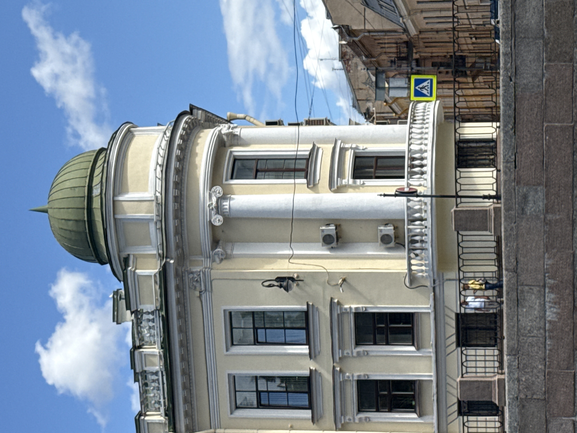 The image shows a view of a building with a green dome in Saint Petersburg, Russia. The building is painted yellow and features classical architectural elements such as columns and ornate moldings. A white balcony with a balustrade wraps around a curved section of the building. Two people are visible standing behind a black metal fence in front of the building. A pedestrian crossing sign is mounted on a pole to the right of the building. The sky is blue with scattered white clouds. The foreground shows a stone embankment. The overall scene depicts a street view of an architecturally significant building in an urban environment.