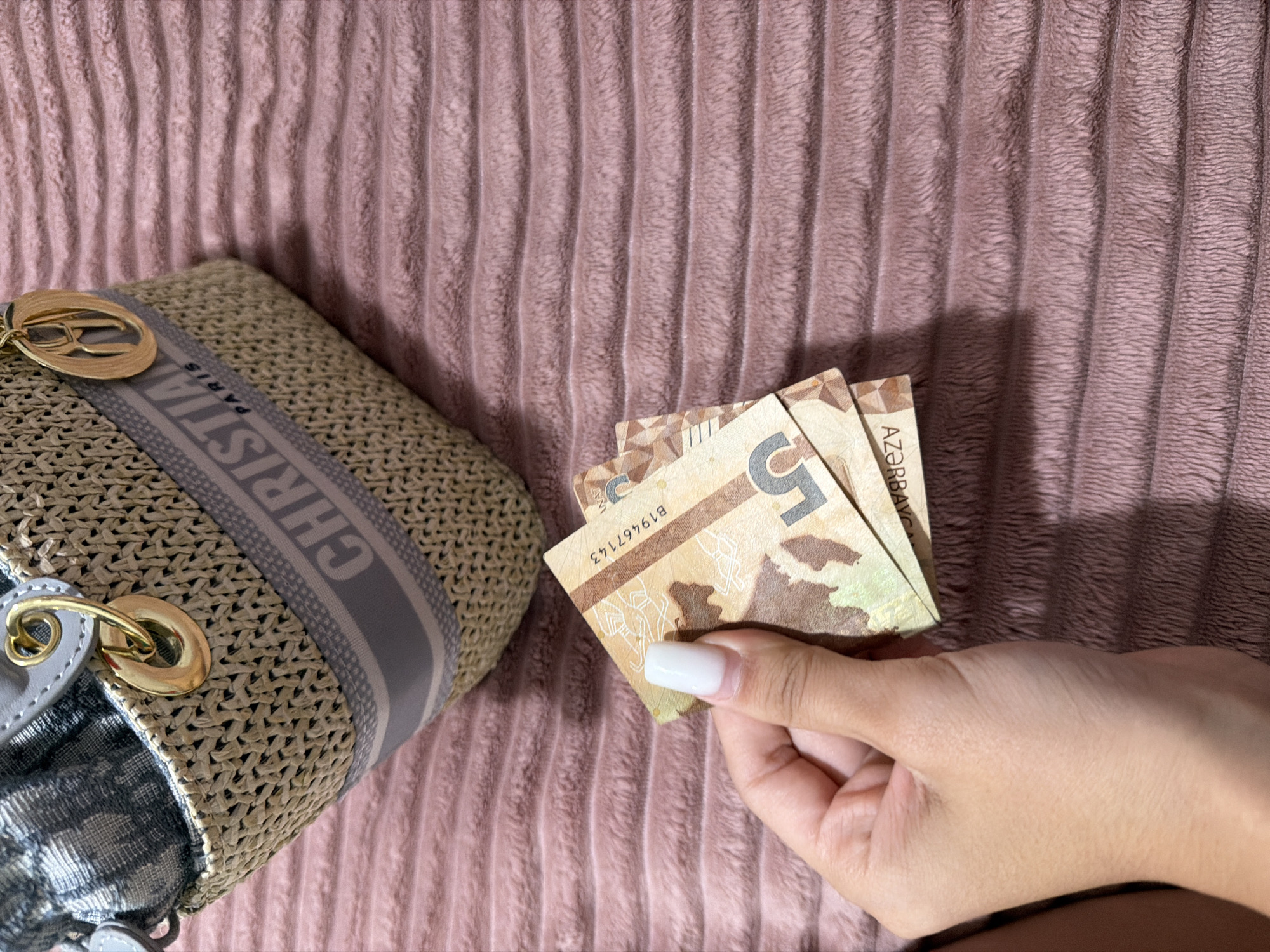In Digah, Azerbaijan, a hand with a white fingernail holds two Azerbaijani 5 manat banknotes. The banknotes are partially obscured by the hand and are fanned out. Behind the hand and banknotes is a close-up view of a beige woven handbag with a grey and white logo band that reads "CHRISTIAN PARIS". The handbag is placed on a pink, ribbed, plush fabric surface.