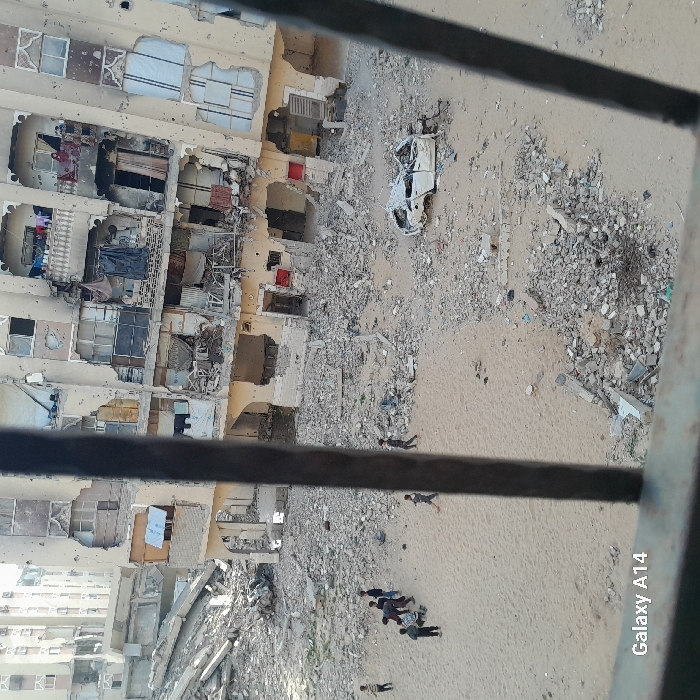 The image, taken from a high vantage point looking down, depicts a scene of significant urban destruction and debris, with several people on the ground. The image is rotated 90 degrees clockwise, so that a tall building appears horizontally oriented on the left side, and the ground extends upwards from the bottom.
On the left side of the frame, there is a multi-story building, heavily damaged. Its facade is pockmarked with holes and collapsed sections, revealing the interiors of multiple apartments. Windows are shattered or missing, and some openings are boarded up or covered with makeshift materials like tarpaulins or fabrics. Clothes lines with laundry are visible hanging from some of the exposed balconies or window openings, suggesting recent or current habitation despite the damage. The building's original color appears to be a light beige or cream. Further to the left and slightly below this main damaged building, parts of other, less damaged buildings can be seen, hinting at a wider urban area.
The ground area, which occupies the majority of the right side and center of the frame, is a mix of sand and extensive rubble. Large piles of concrete chunks, twisted rebar, and assorted debris are scattered across the sandy ground. In the upper central part of the ground area, a heavily damaged and burnt-out white car wreck is visible, appearing as little more than a chassis with missing doors and windows.
Several small groups and individual figures, likely people, are present on the sandy ground amidst the rubble. Towards the lower right corner, a cluster of approximately five to six individuals is gathered. They appear to be standing or kneeling, some looking down at the ground or at something among them. Further up and to the right from this group, two more individuals are seen, one lying face down and another lying on their side. Near the bottom edge of the frame, another lone figure is visible, also lying on the ground. Another individual is standing alone closer to the damaged building on the left. The figures are too distant to discern specific features or emotions, but their posture suggests a somber or observant mood.
The scene is lit by natural daylight, indicating it is daytime. The overall impression is one of devastation and hardship. In the very foreground, a dark horizontal bar runs across the image, suggesting the photo was taken through a window, from a balcony, or another elevated structure, with the bar being a railing or frame component.
In the bottom right corner of the original image, faint white text reads "Galaxy A14", indicating the device used to capture the photo.