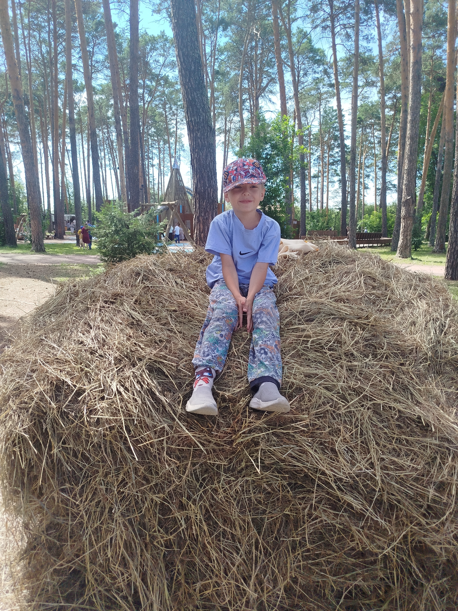 A young boy wearing a blue t-shirt, patterned pants, and a colorful baseball cap sits on a large haystack. He is looking at the camera and smiling. The background consists of a forest of tall, slender pine trees under a bright, partly cloudy sky. In the distance, there are some wooden structures and a few other people can be seen. The ground is covered in dirt and scattered pine needles. The image appears to have been taken outdoors during the daytime. The location is the city of Zavodoukovsk, Russia.