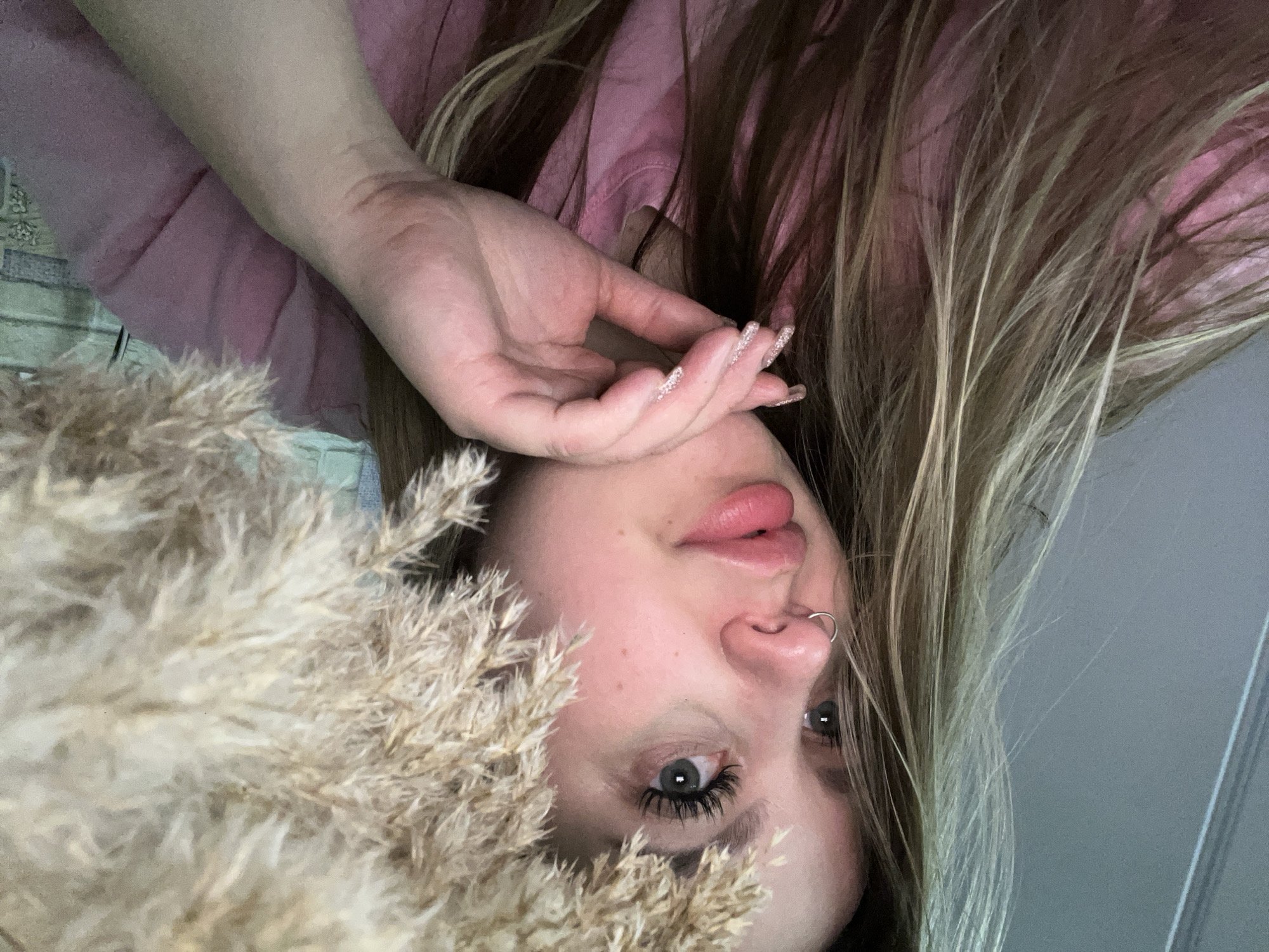 A woman with blonde hair is pictured in a close-up shot. She is lying on her back, with her head facing the bottom of the frame. Her left hand is positioned near her face, with her fingers slightly spread apart. She has a nose ring and noticeable eyelashes. Her gaze is directed towards the viewer. A bunch of beige, fluffy grass-like material is prominently positioned in the lower-left area of the picture. The woman is wearing a pink top. Part of a light green and patterned fabric is also visible. The background has a grey wall.
This image was not taken in Port-Katon, Russia. It contains no references to suggest it was.