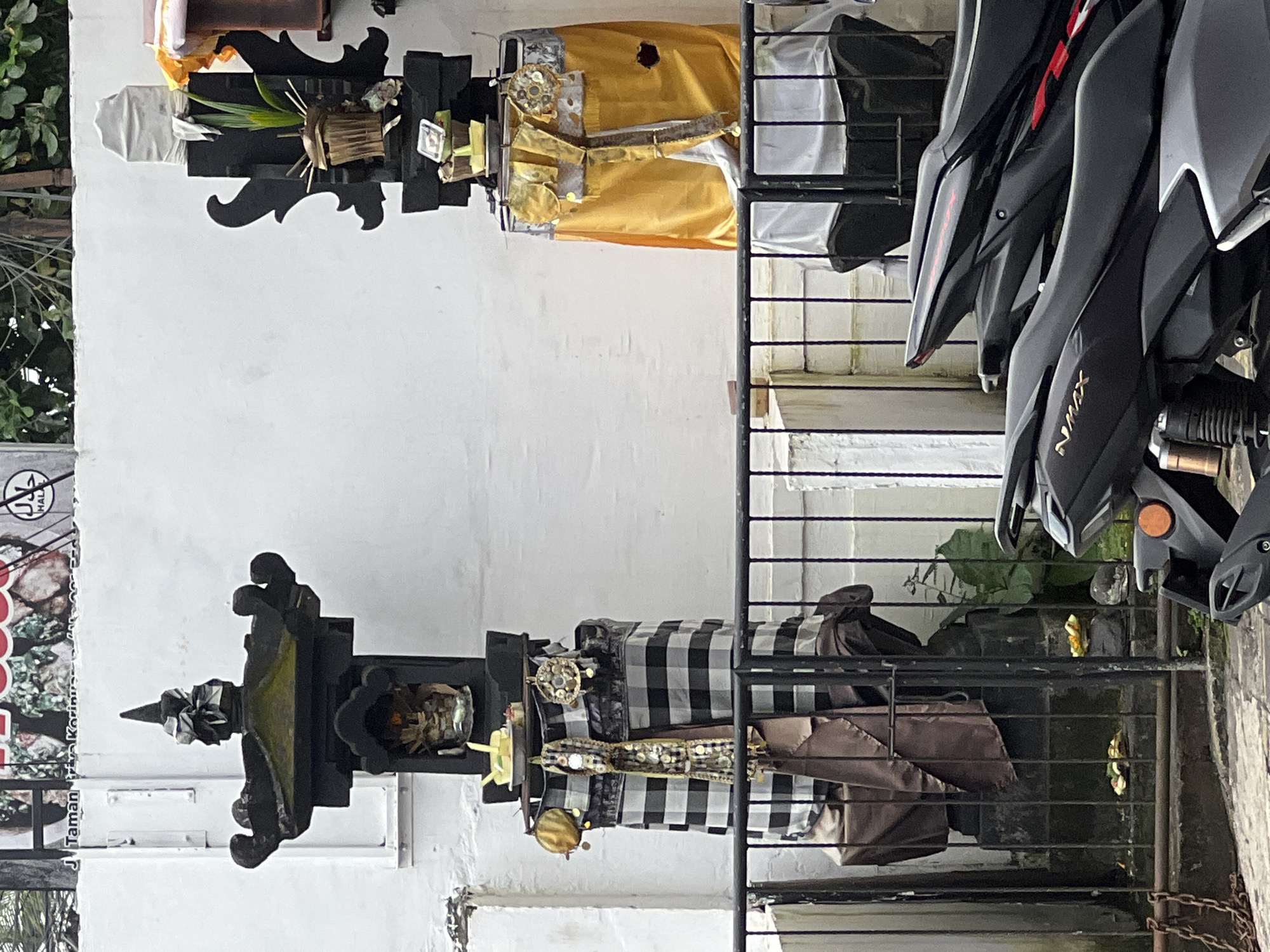 The image captures an outdoor scene in Kuta Selatan, Indonesia, depicting a blend of traditional Balinese religious offerings and modern street elements. The photo appears to be taken during the daytime under bright, possibly overcast, conditions.
In the left half of the frame, two traditional Balinese shrines or offering stands are prominently displayed against a plain white wall. The shrine on the left is adorned with a black and white checkered "poleng" cloth, a common pattern in Balinese Hinduism, and features intricate offerings including woven baskets, green foliage, and a golden decorative object. The adjacent shrine to its right is draped in a vibrant yellow cloth, also holding various offerings and decorative elements.
A black metal fence or railing separates these sacred installations from the right side of the image, where several dark-colored motorcycles are parked. At least two of these are identifiable as Yamaha NMAX scooters, with the "NMAX" branding visible in gold lettering on their side panels. The motorcycles are parked closely together, suggesting a street or designated parking area.
To the far left, a fragment of a commercial sign is visible, featuring a "HALAL" logo and partially legible text such as "J. Taman P..." and "Korinu...". Green foliage can be seen in the background behind the fence and shrines. No people are visible, and the scene depicts static objects.