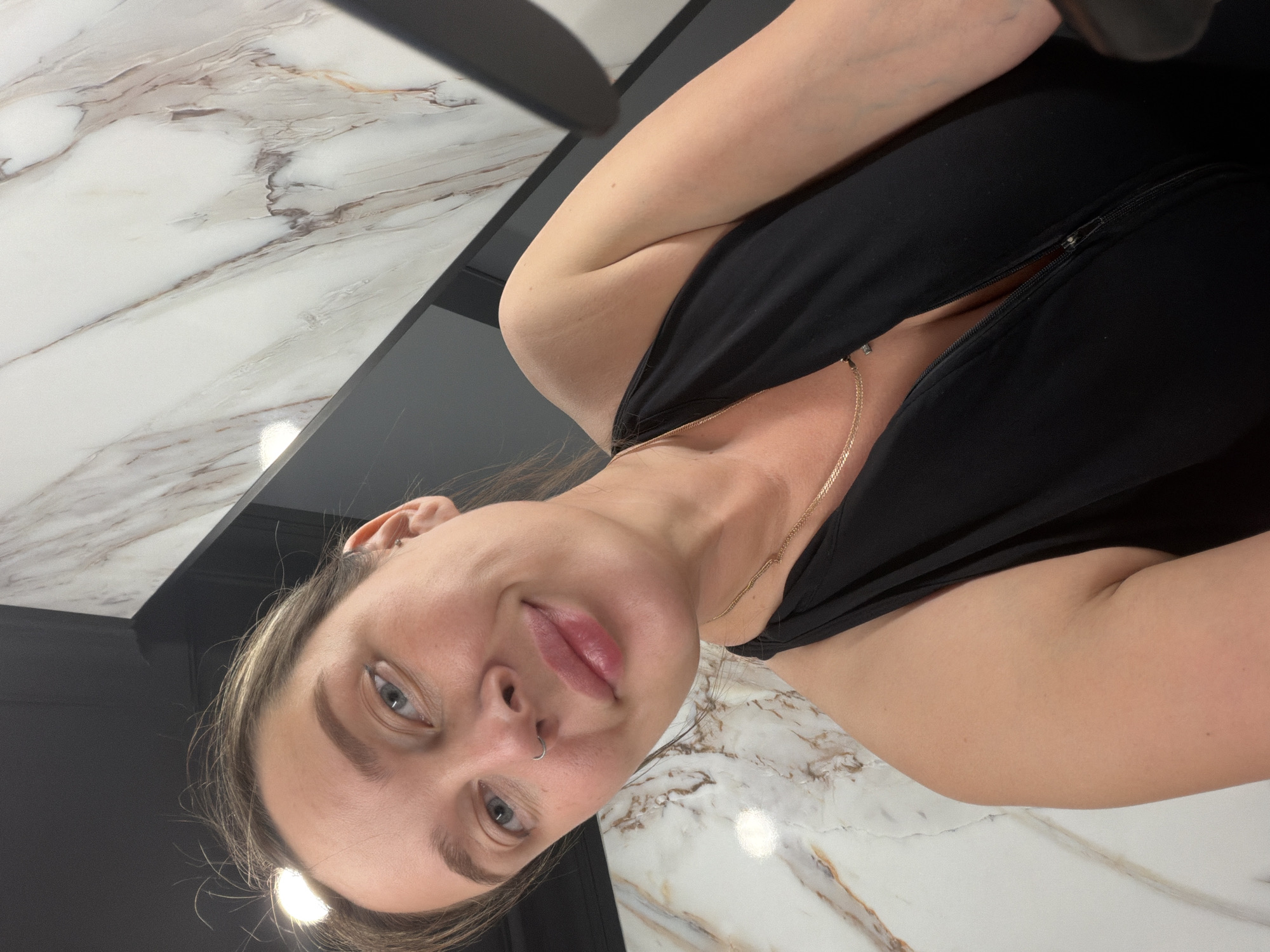 A woman is posing for a selfie. She has fair skin, light-colored eyes, and brown hair with blonde highlights. She is wearing a black top and a gold necklace. She has a nose ring. The background includes a marble countertop and a black wall or cabinet. The location is not identifiable as Moscow, Russia.