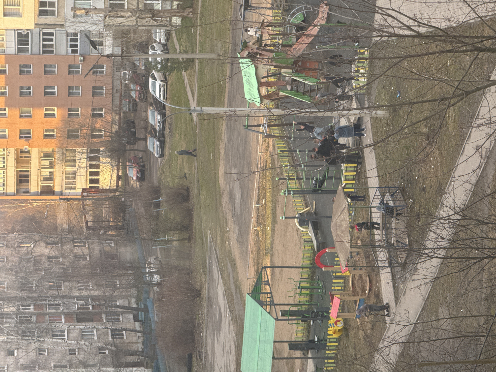 An elevated, oblique-angle view depicts a residential area in Kladovitsy, Russia, with apartment buildings to the left. A playground is prominent in the foreground and midground, featuring slides, climbing structures, and covered play areas. Bare trees are interspersed throughout the scene. Several individuals, including adults and children, are present on the playground. Some are standing and interacting near the climbing structures, while others are dispersed throughout the area. Parked cars are visible on a street between the playground and the apartment buildings. The environment suggests a late fall or early spring setting.