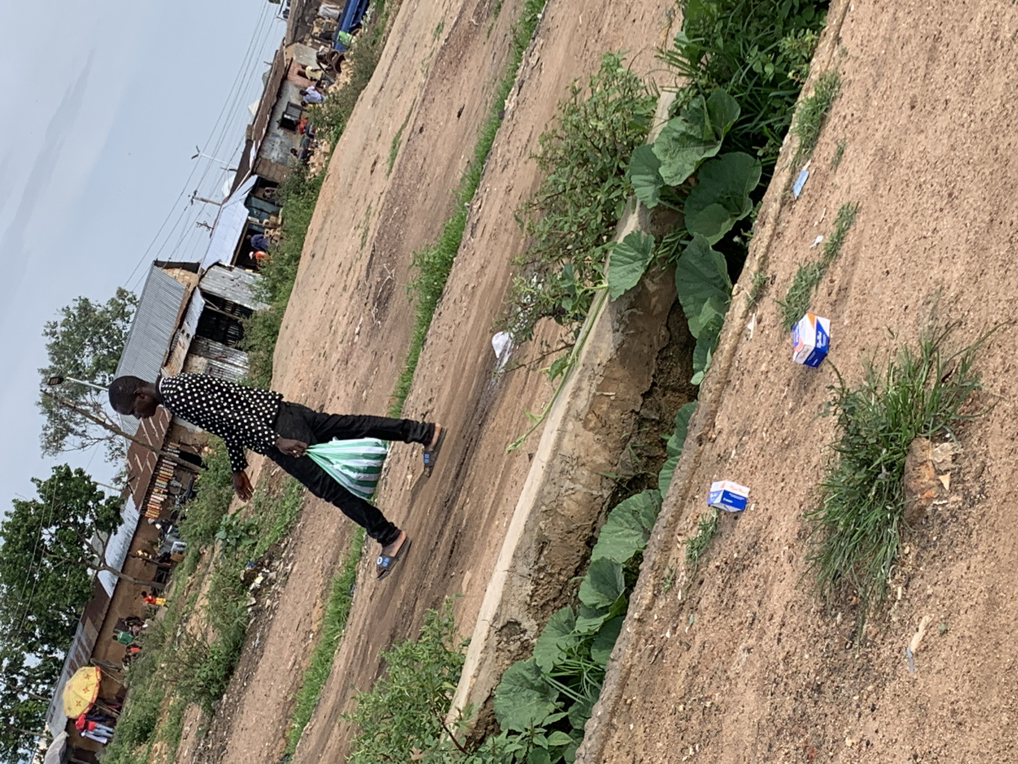 The image depicts a scene in Bukuru, Nigeria, with a person walking on a dirt path next to a drainage ditch filled with lush green vegetation. The person, dressed in a black and white polka dot shirt and black trousers, is carrying a green and white striped bag over their shoulder. They are wearing sandals.
The setting appears to be a semi-urban or rural area, with makeshift structures and shops visible in the background. Some of these structures have corrugated metal roofs, suggesting a market or informal settlement. People are milling around these buildings, and there is a yellow umbrella visible, possibly indicating a vendor or a stall. The ground is predominantly dirt, with patches of grass growing along the sides of the path and in the ditch. There are also some discarded items, including small white and blue boxes, and white paper scattered on the ground.
The time of day is difficult to ascertain precisely, but the lighting suggests it could be daytime, perhaps mid-morning or afternoon, with overcast skies contributing to the soft light. The overall atmosphere seems ordinary, with the individual engaged in a mundane activity of walking through the area. The presence of the ditch and surrounding vegetation adds a natural element to the otherwise built environment. No specific event is clearly depicted, but the presence of multiple people in the background suggests a bustling local area.
