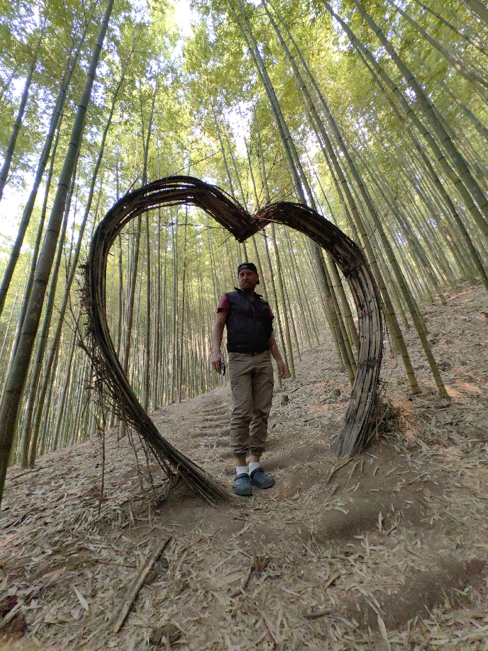 A male subject stands centrally within a large, heart-shaped structure crafted from intertwined dark branches, situated on a sloped path within a dense bamboo forest in Mù Cang Chải, Vietnam. The man is Caucasian, with a beard and a black baseball cap worn backward. He wears a dark-colored sleeveless vest over a darker short-sleeved shirt, tan cargo pants cuffed at the ankles, white socks, and dark blue or grey clogs. His right hand holds a small, dark rectangular object, possibly a phone or camera, while his left arm hangs by his side. He looks directly towards the viewer. The ground beneath him and extending throughout the foreground is dry, reddish-brown earth covered with numerous fallen light brown bamboo leaves and debris. Subtle steps are visible carved into the sloped terrain behind the subject. Numerous tall, slender green bamboo stalks rise steeply into the bright, filtered sunlight overhead, forming a dense canopy and dominating the background.
