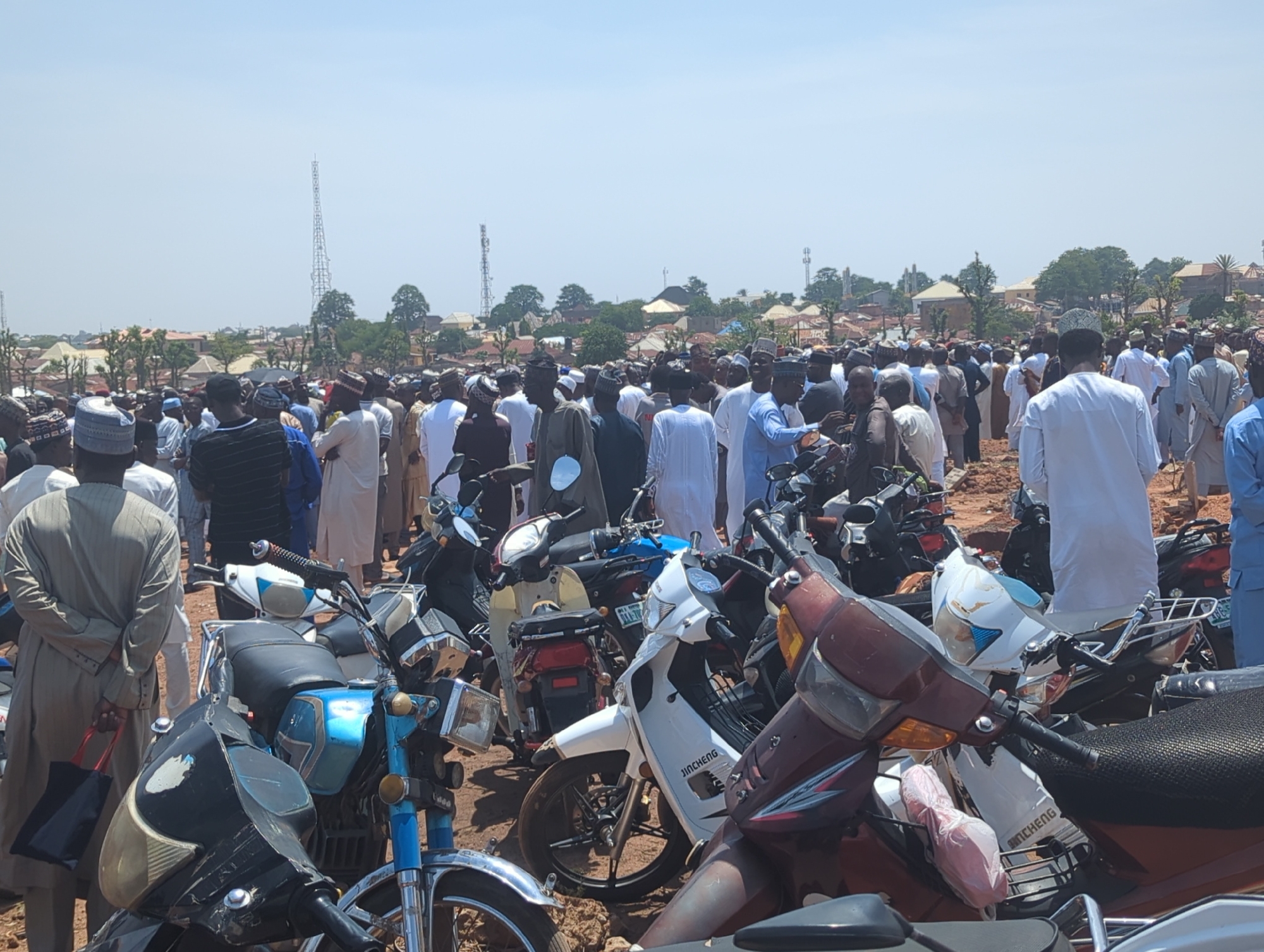 This outdoor photograph captures a large gathering of men in what appears to be a communal event. In the foreground, numerous motorcycles and scooters are parked densely, partially obscuring the crowd behind them. One white scooter prominently displays "JINCHENG" on its side.
The majority of the men in the crowd are dressed in traditional attire, including long robes and various types of caps, suggesting a cultural or religious context for the assembly. They are densely packed together, facing generally in the same direction, implying a central point of focus or activity not visible within the frame.
The setting is an open, unpaved area, likely dirt. In the mid-ground and background, a sprawling settlement of buildings, likely houses, is visible under a clear, bright blue sky. Several tall, metal communication towers or antennae rise above the distant structures and trees. The clear sky and strong illumination indicate it is daytime, likely morning or early afternoon. The scene evokes a bustling community event in a semi-urban or town environment, consistent with a location like Sardauna, Nigeria. The overall mood appears orderly and composed.
