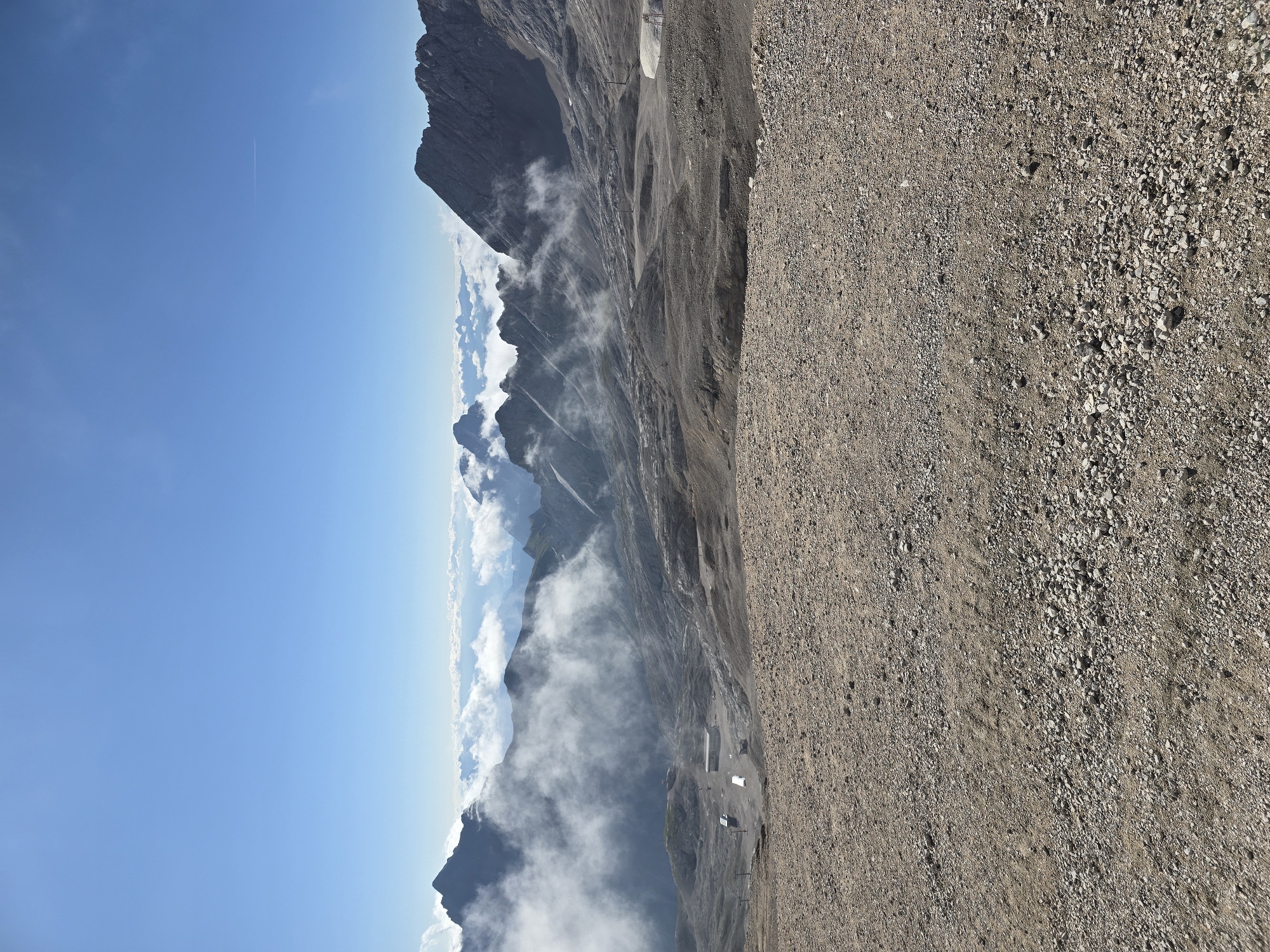 This is a scenic, eye-level, wide-angle shot of a mountainous landscape, likely taken on a sunny day. In the foreground, a barren, gravelly ground stretches across the frame. Beyond this, the terrain slopes downwards, revealing a valley dotted with several buildings and structures, possibly a small village or a resort area. Wisps of white clouds drift through the valley and around the surrounding mountain peaks. The mountains themselves are rugged and rocky, with some peaks shrouded in mist. The sky is a clear, bright blue, with a faint contrail visible in the upper right quadrant. The image appears to be of the Zugspitze area, Germany's highest peak, located near Garmisch-Partenkirchen. There are no people or animals visible in the image.
