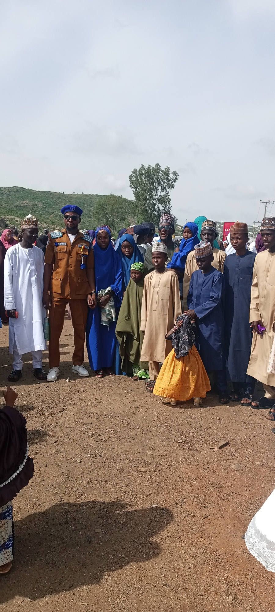 A group of people stands on a dirt ground in what appears to be an outdoor setting. The sky is overcast and a hill with sparse vegetation is visible in the background, suggesting a location near or in a semi-arid region.
The individuals in the group are dressed in traditional Muslim attire. There are males and females, including children. Their clothing colors vary. A man wearing a brown uniform with a blue hat stands out among the group, with what appears to be his family around him.
The ground appears to be soil.
Based on the information and clues, this photograph appears to be taken in Naraguta Hausawa, Nigeria.