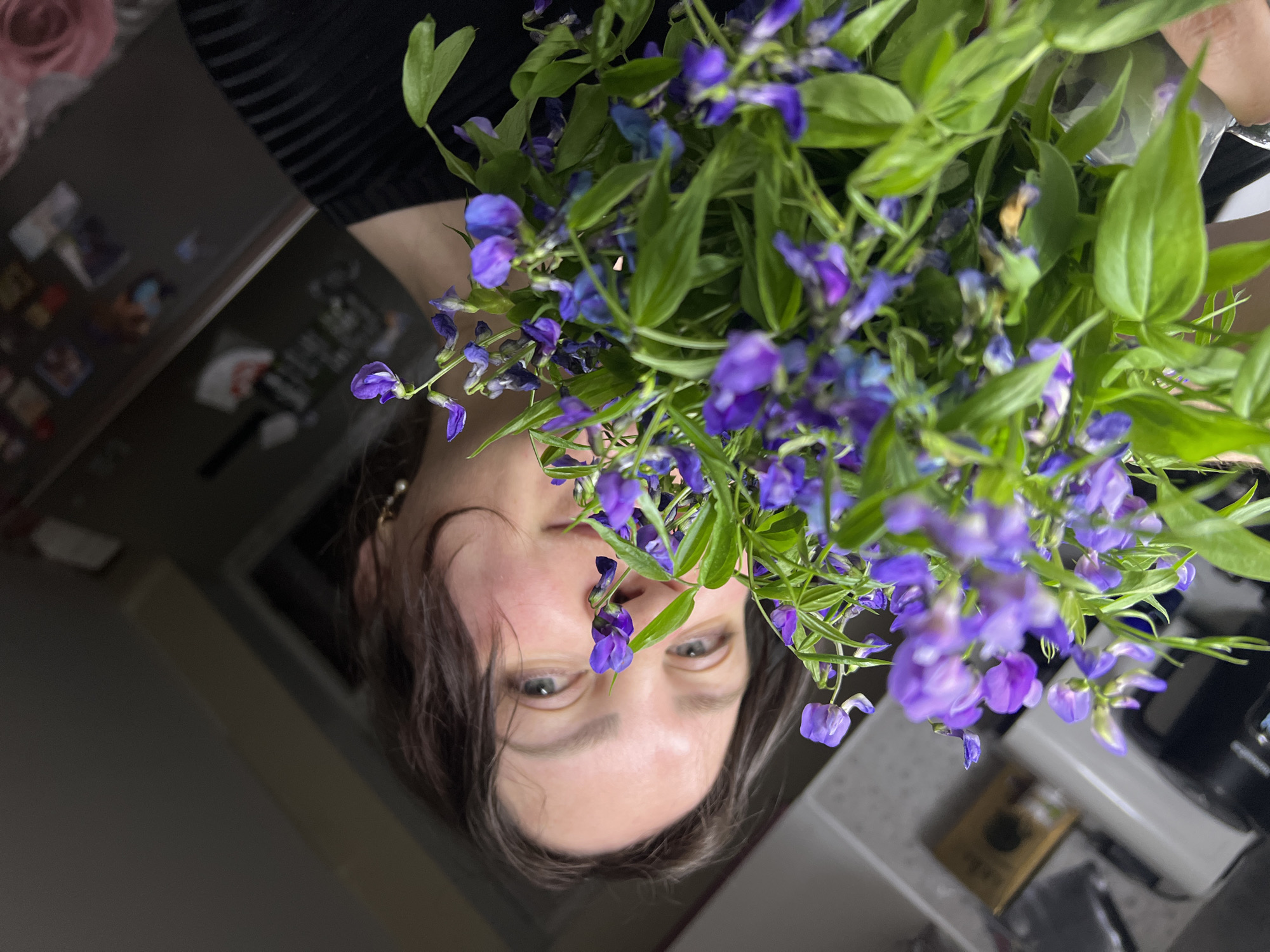 A woman holds a bouquet of purple flowers close to her face. The photograph is taken from an upside-down perspective. The background includes elements of a kitchen setting, such as countertops, cabinets, and other household objects. Noviy, Russia is not indicated.