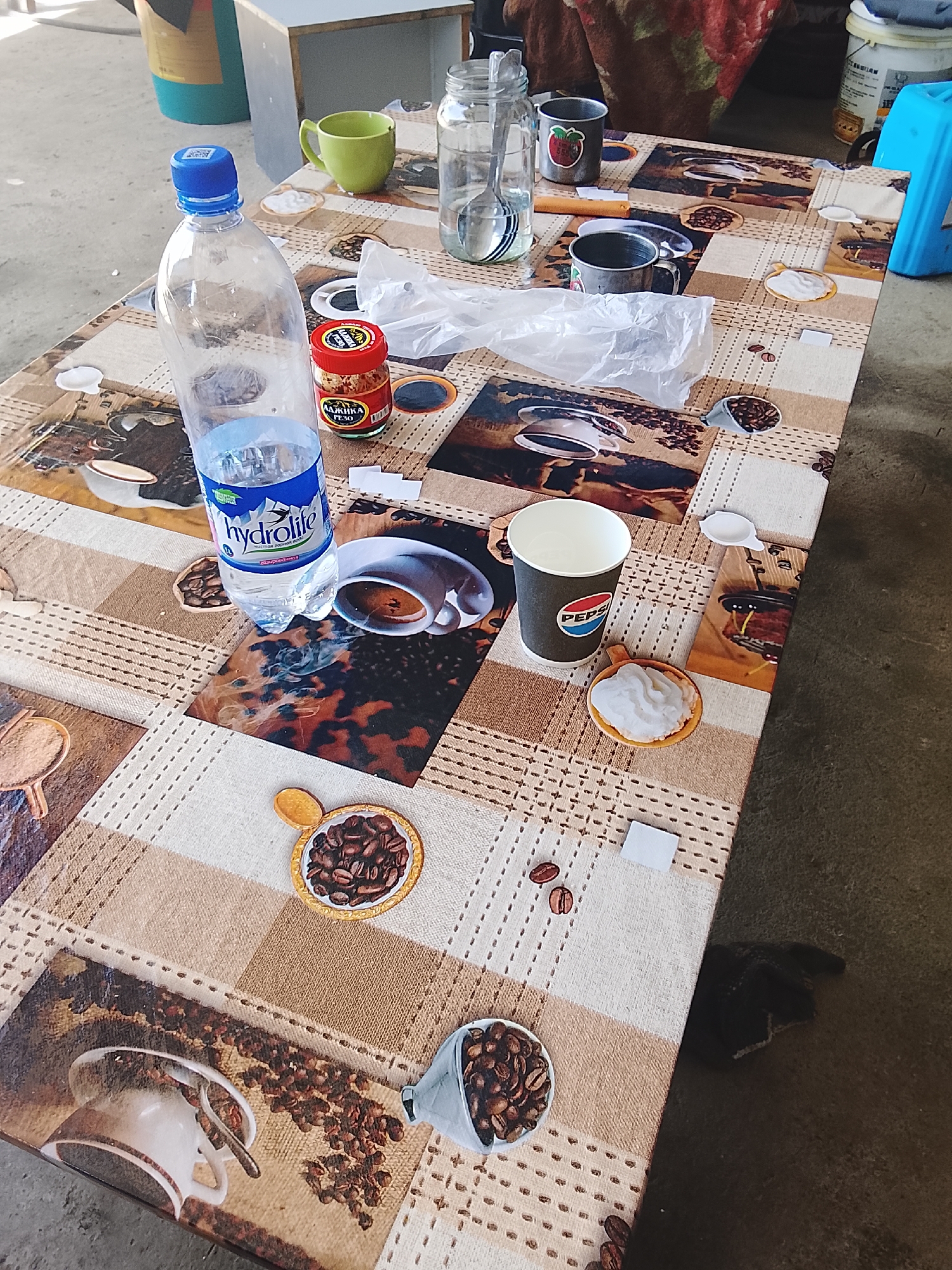 A table covered with a coffee-themed cloth is set with various items. In the foreground, a clear plastic bottle of "Hydrolite" mineral water stands next to a small red jar of what appears to be spices or seasoning, labeled "ЛАЙМКА REJO". To the right of the water bottle, a dark gray Pepsi paper cup is positioned next to a small ceramic dish filled with a white substance, possibly cream or yogurt. Further back on the table, a glass jar contains a spoon and water, a green mug, and two metal mugs, one with a strawberry sticker. The table itself is decorated with a collage of images featuring coffee cups and beans. The background shows a blurred environment, possibly outdoors or in a garage, with a blue plastic stool and containers visible. The scene suggests a casual setting, perhaps a break or a meal.