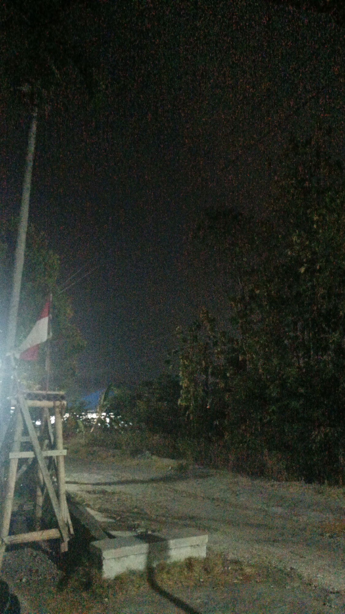 A nighttime outdoor scene in Sambi, Indonesia, features a dirt path with some scattered concrete blocks. On the left side of the path, a wooden structure resembling a scaffold or a partially built platform is visible. A flag with red and white horizontal stripes, likely the Indonesian flag, is displayed on a pole next to the structure. Trees and foliage form a dense backdrop to the right of the path. The lighting is dim, suggesting artificial illumination is present, possibly from a street lamp or nearby buildings, casting shadows on the ground. The overall atmosphere is dark and somewhat rural.