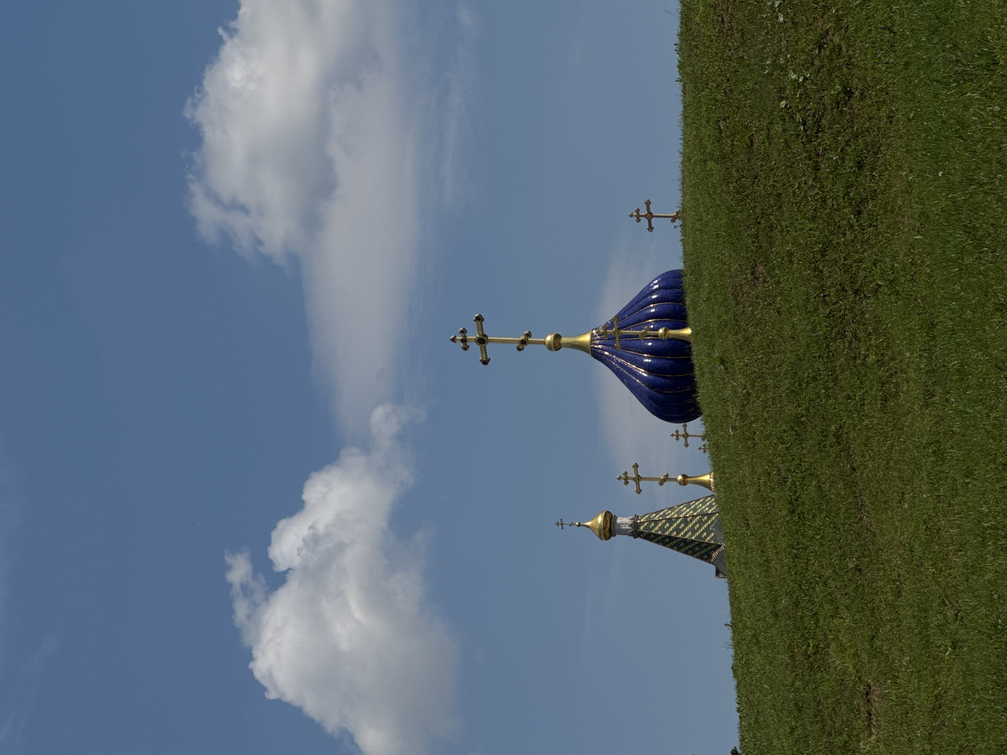 This image captures a vibrant outdoor scene featuring elements of Russian Orthodox architecture against a bright sky. The perspective is from a slightly low angle, looking up towards the structures.
In the foreground, covering the right side of the frame, is a well-maintained, lush green grassy slope, suggesting a hill or manicured lawn. The sky, a clear blue, occupies the left and upper portions of the image, dotted with several soft, white clouds.
Two distinct church domes (cupolas) are visible on the left side, rising above the grass line. The most prominent dome is a deep, rich blue, possibly made of glazed tiles or mosaic, with intricate vertical ribbing highlighted in gold or white. It is topped with a golden Orthodox cross, characterized by its three horizontal bars and an additional diagonal bar at the bottom. A second, smaller dome is partially visible behind and to the left of the main blue one. This dome appears to be patterned in green and gold, also surmounted by a golden Orthodox cross.
Several other golden Orthodox crosses are scattered across the blue sky, some seemingly detached from visible structures, indicating other nearby architectural elements that are out of frame or more distant.
The scene depicts a peaceful, sunny day. No people are visible, and there is no discernible activity taking place. The architectural style, particularly the onion domes and Orthodox crosses, strongly suggests a setting in Moscow, Russia, consistent with the provided context. There is no visible text in the image.