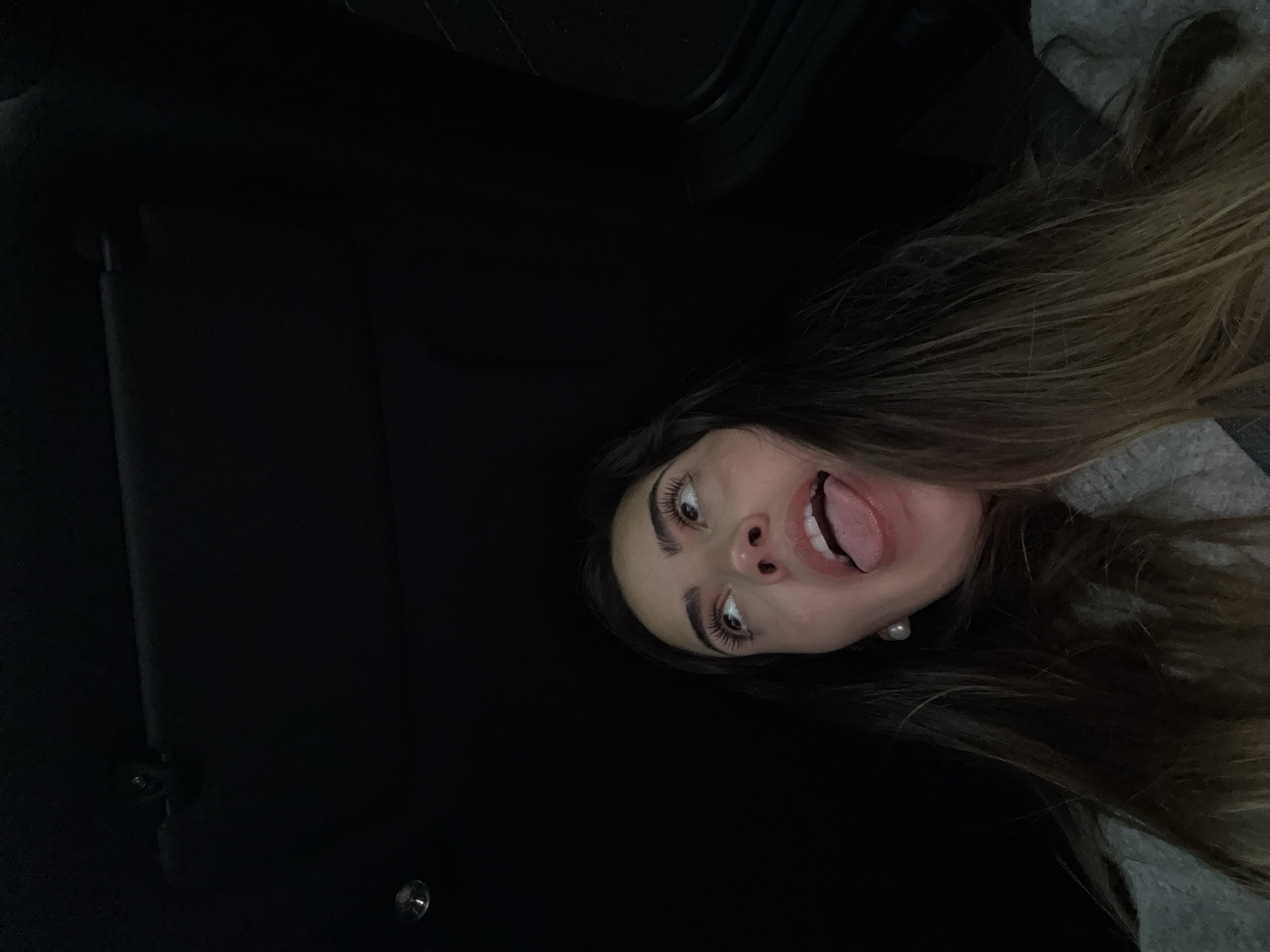 A woman with long brown hair is posing with her mouth open and tongue extended. She is wearing a light gray top and a pearl earring is visible on her left ear. She is in a car with black interior. The image doesn't provide information to determine if the photograph was taken in the city of San Bernardino Tlaxcalancingo, Mexico.