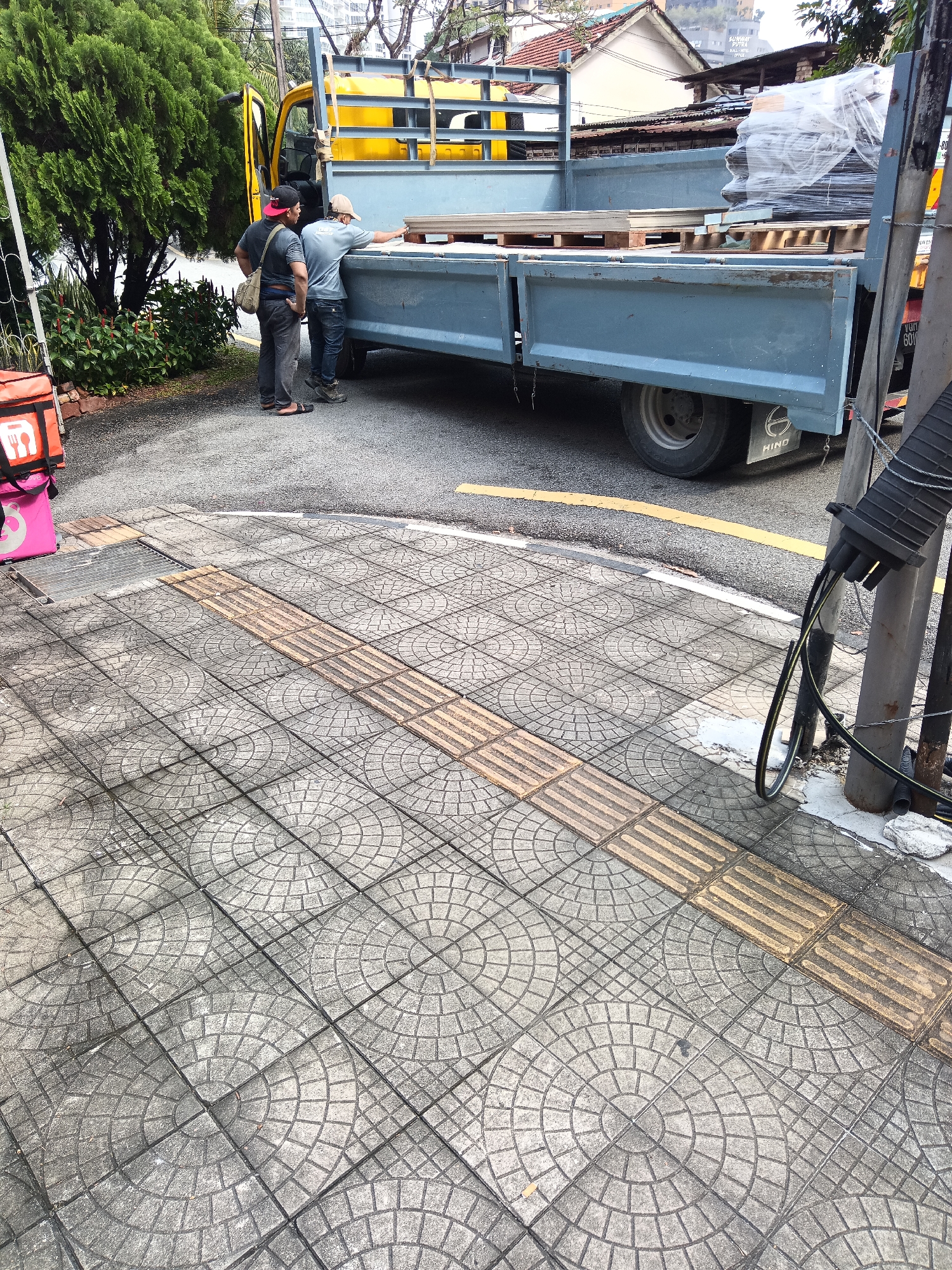 In Kuala Lumpur, Malaysia, two men stand near the rear of a blue flatbed truck, which is loaded with wooden pallets covered in plastic wrap. The truck is parked on a paved road adjacent to a sidewalk. The sidewalk is made of grey concrete tiles with a circular pattern and features a tactile paving strip. To the left of the sidewalk, a pink food delivery bag is visible. Behind the truck, a yellow truck is partially visible, along with trees and buildings. A metal pole with cables is attached to the sidewalk.