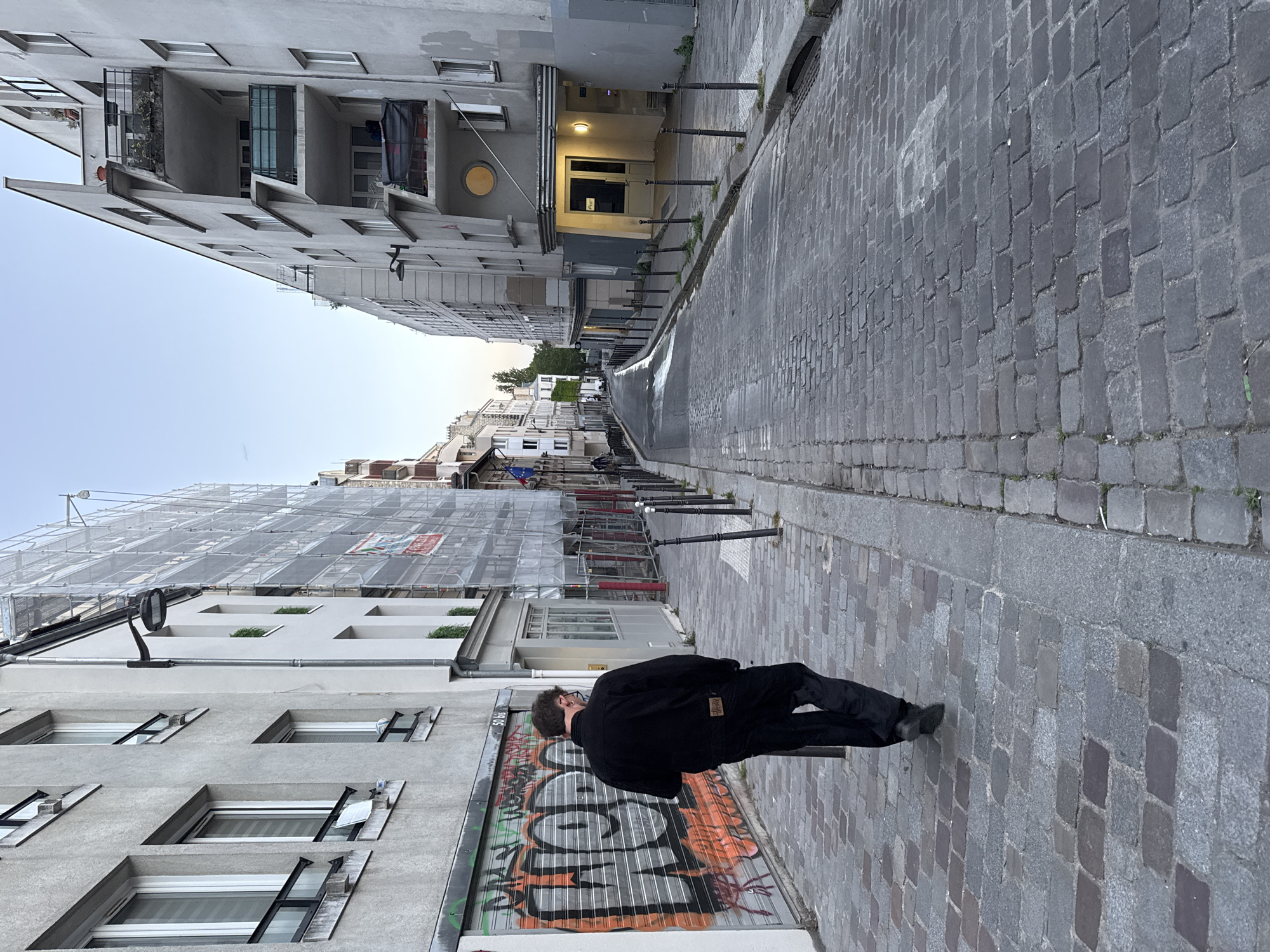 The image captures a perspective of a narrow, cobblestone street in Paris, France, lined with tall, grey buildings under a clear sky. A lone individual, dressed in dark clothing including a black jacket and pants, walks away from the camera down the center of the street.
On the left side of the street in the foreground, a building features a metal rolling shutter heavily covered in colorful, stylized graffiti. Further down this side, a significant portion of another building is enveloped in white scaffolding, indicating ongoing construction or maintenance. This building exhibits distinct architectural details, including angular, triangular protrusions above some windows.
The right side of the street is bordered by a series of short, black bollards. The buildings on this side are also multi-storied and grey, featuring various windows and balconies, some adorned with small plants. One building entrance further along is illuminated by an outdoor light fixture, casting a warm glow on the doorway. A distinctive circular window is visible on a higher floor of one of these buildings.
The street surface is paved with dark grey cobblestones, showing different shades and textures. The clear, bright sky suggests it is daytime, with a soft, ambient light illuminating the scene. While there is graffiti on the shutter and a red and white sign on the scaffolding, no specific, legible text can be discerned. The overall impression is that of a quiet, urban street scene.