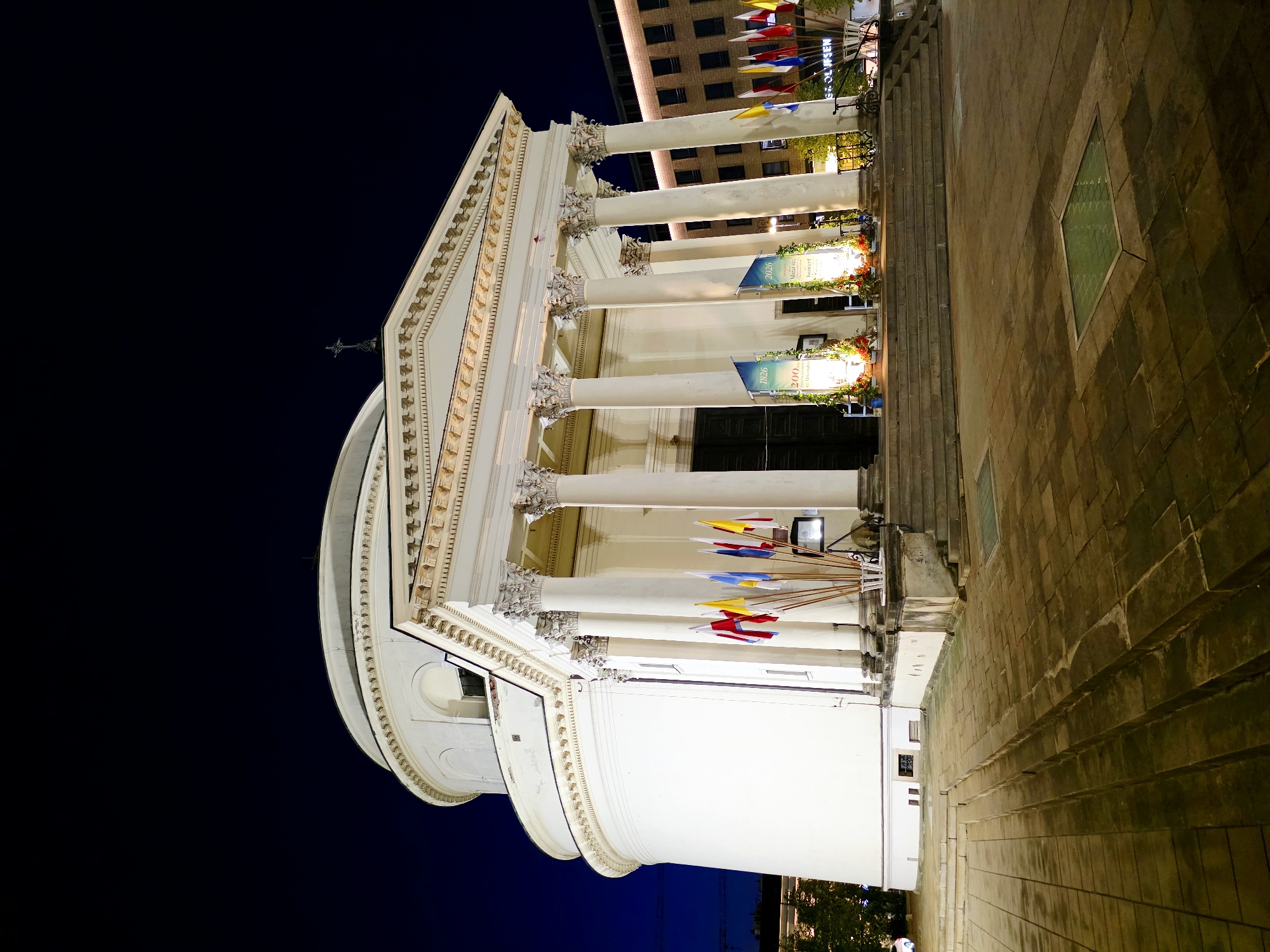 The image shows the exterior of the Holy Trinity Church in Warsaw, Poland, at night. The church, a neoclassical building, is illuminated, highlighting its white facade, prominent columns, and domed roof. Several flags are displayed in front of the church, and two banners are hung near the entrance. The surrounding area is paved with stone, and the sky is dark blue. In the background, modern buildings are visible, also lit up. The scene appears to be part of a special occasion or commemoration, suggested by the banners and flags.