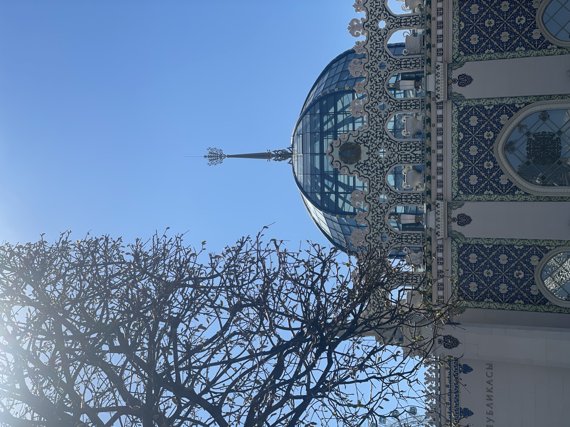 The image captures a view of the building's architecture in Moscow, Russia, featuring a glass dome with an ornate spire atop the structure, set against a clear, blue sky. The building's facade is adorned with intricate blue and white patterned tiles, arched windows, and decorative elements. A bare tree, with only a few leaves budding, is prominently featured in the foreground, partially obscuring the lower portion of the building. The photo appears to have been taken during the daytime.