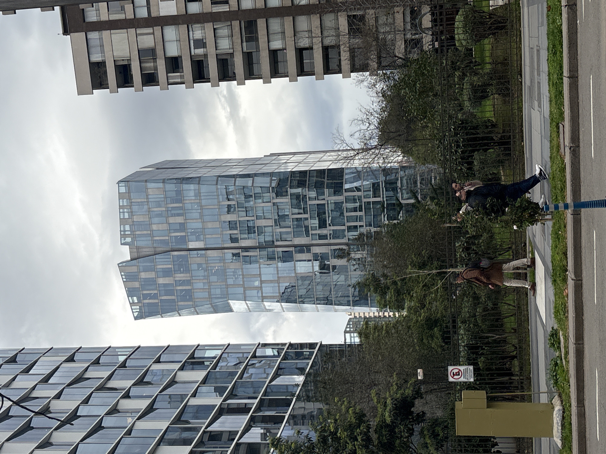 The image captures a street-level view of modern architecture in Santiago, Chile, on an overcast day. In the foreground, a sidewalk and street are visible, with a yellow traffic signal pole on the left. Two pedestrians are walking on the sidewalk. The first, on the left, is a man wearing a brown coat and light-colored pants, carrying a backpack. The second, further to the right, is a man wearing a dark jacket and dark pants, also with a backpack.
Behind the pedestrians, a dark metal fence encloses a green space with trees and bushes. In the background, three prominent buildings dominate the skyline. The tallest, in the center, is a distinctive glass skyscraper with a unique, warped facade. To its left, another tall, rectangular glass skyscraper with a grid-like pattern of windows is partially visible. To the right of the central skyscraper, a narrower, older-looking building with a beige facade and numerous balconies stands tall. The sky is heavily clouded. The scene suggests an urban environment with a mix of contemporary and older architectural styles.