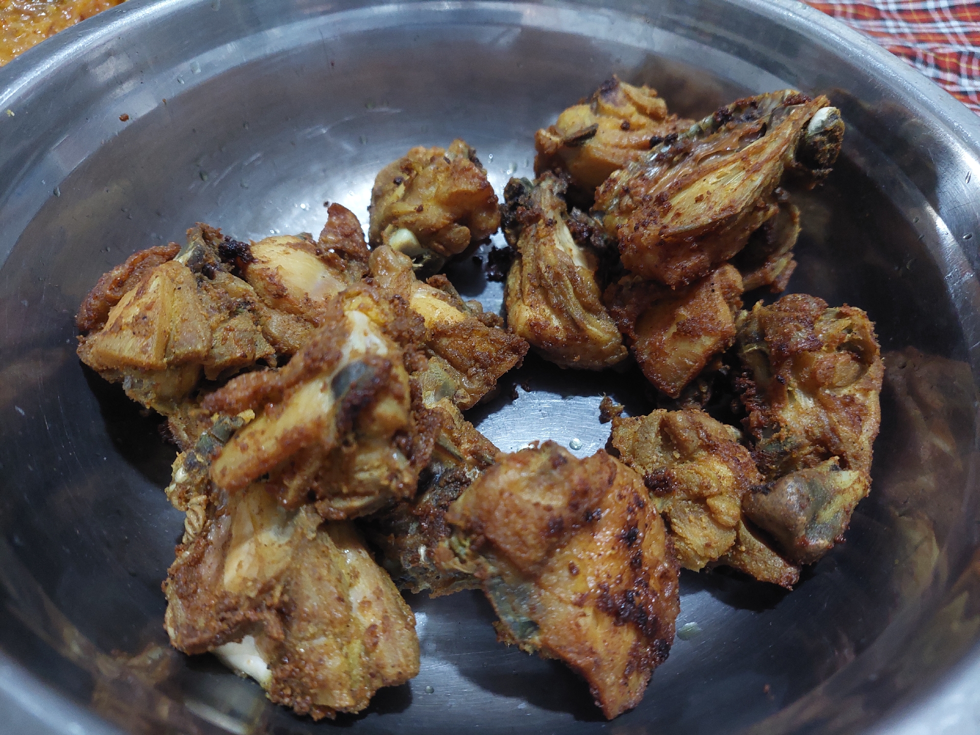 A close-up, overhead shot showcases a metal bowl filled with an assortment of fried chicken pieces. The chicken appears to be seasoned and cooked to a golden-brown hue, with some pieces exhibiting a darker, crispier exterior. The bowl itself is metallic and appears to be a serving dish. In the background, a portion of a red and white checkered cloth is visible, suggesting a casual dining setting. The scene is well-lit, highlighting the texture and color of the fried chicken. The image provides no context for the specific location within the city of Jelutung, Indonesia, as no identifiable landmarks or specific environmental cues are present.