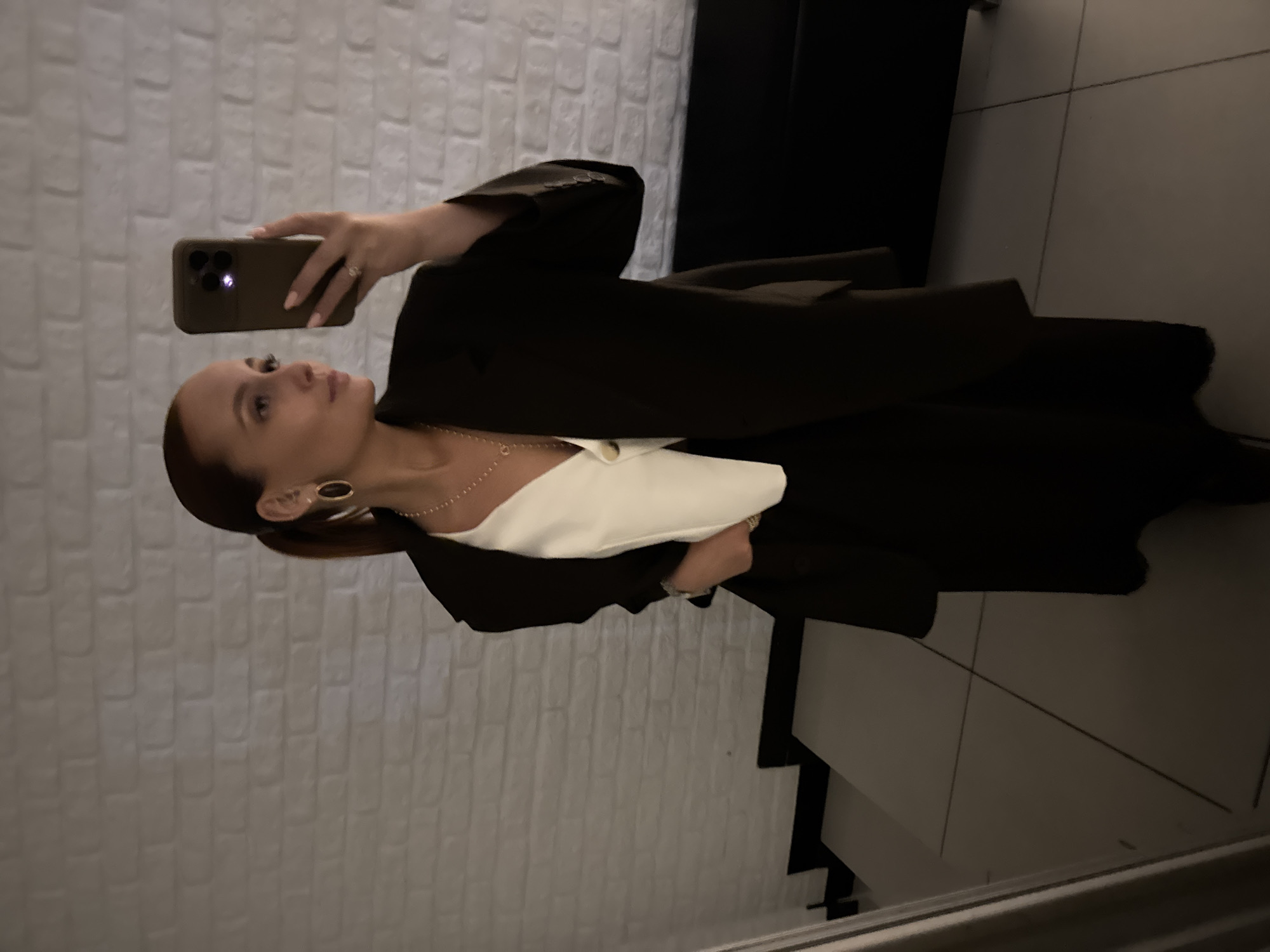 A woman is taking a selfie with her cell phone. She has red hair pulled back in a ponytail, gold hoop earrings, and is wearing a black blazer over a white top with a gold necklace. She appears to be indoors with a white brick wall behind her. Next to the wall is a staircase with dark steps. No additional location information.