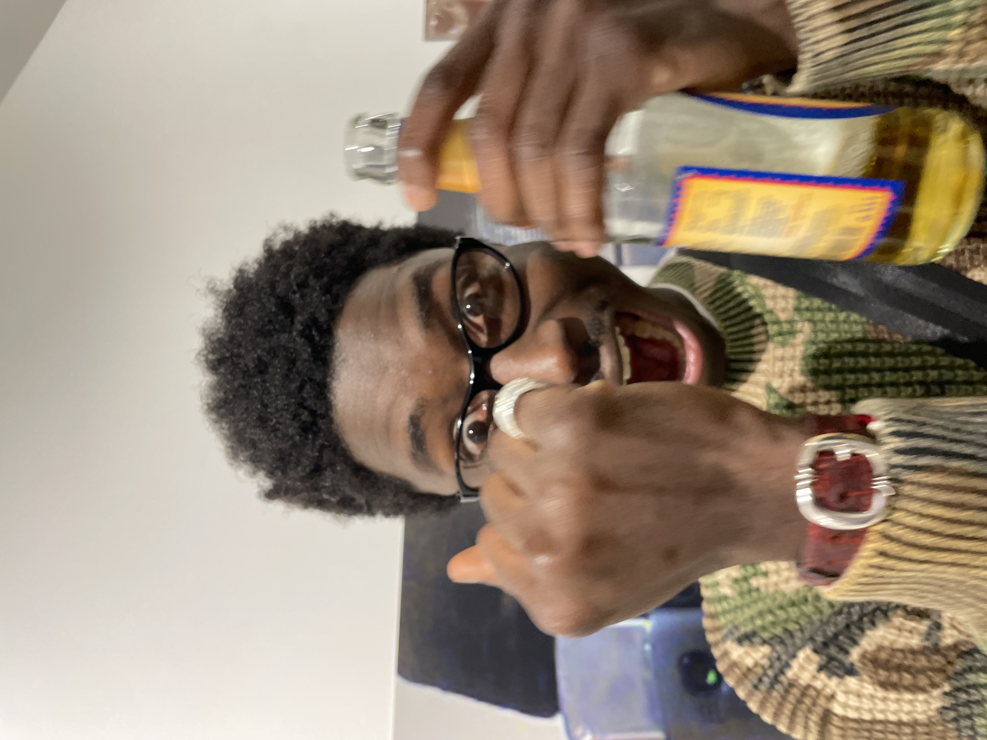 This close-up image depicts an individual in an indoor setting located in Paris, France. The central figure is a person with dark skin, short curly black hair, a mustache, and black-framed glasses. They face the camera directly, exhibiting wide eyes and an open mouth, conveying an animated expression. They wear a knitted sweater featuring a camouflage-like pattern in shades of brown, green, and beige. A dark strap traverses their torso. The individual holds an open clear glass bottle, approximately half-filled with a yellow-gold liquid, in their right hand. Their left hand is raised, with the index finger slightly curled and displaying a silver-colored ring with a ridged texture. A watch with a red band and a prominent silver buckle is visible on their left wrist. The background consists predominantly of a plain white wall, with a dark object, possibly upholstered furniture or an art piece, showing blue reflections in the upper-left quadrant.