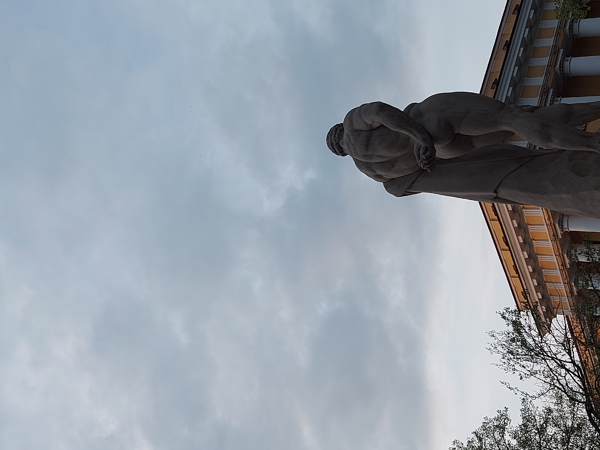 The image shows a low-angle view of a sculpture in Saint Petersburg, Russia. The sculpture appears to be a male figure in a dynamic pose. The sky occupies a large portion of the frame, with soft clouds. The building next to the sculpture has a classical style, with columns and a yellow facade. Bare tree branches are also visible. The location is likely near the State Hermitage Museum, where similar sculptures are located.