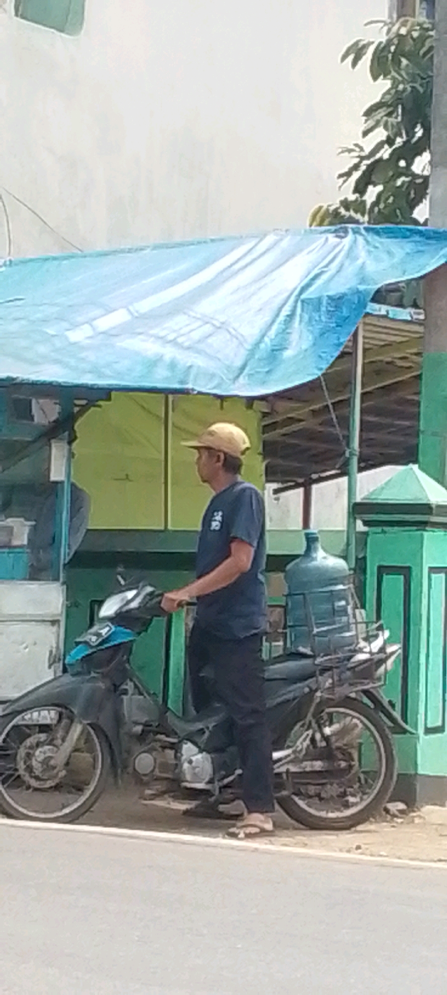 A man wearing a light brown hat and a dark blue t-shirt is standing next to a motorcycle in Cilaku, Indonesia. The motorcycle is black and has a large blue water jug secured to the back. The man is holding onto the handlebars of the motorcycle, and his left foot is on the ground. Behind the man and the motorcycle is a green structure with a blue tarp covering the top, creating a makeshift shelter or stall. The environment appears to be outdoors, possibly a roadside or market area, with a paved surface in the foreground that resembles a road. The background shows a light-colored wall and some greenery, including the leaves of a tree. The lighting suggests it is daytime.