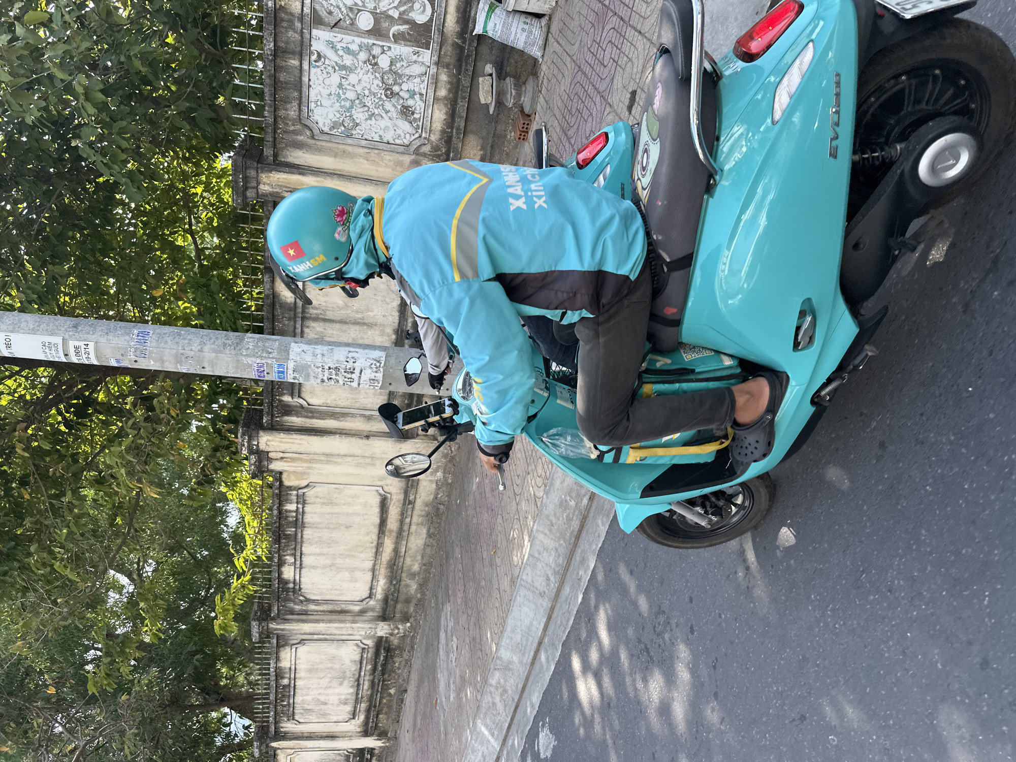 A person wearing a turquoise jacket and helmet is riding a turquoise scooter on a paved road. The scooter has a yellow strap hanging from the back. The rider is wearing dark pants and black crocs. The jacket has text that says "Xanh" and "xinch". The helmet has a Vietnamese flag sticker and a lotus flower sticker. In the background, there is a concrete wall with decorative elements and a street lamp with various signs. Trees are visible behind the wall, and there are some bags and bricks on the sidewalk. The scene appears to be in a city setting, likely in Bac Nha Trang Ward, Vietnam, given the context provided.