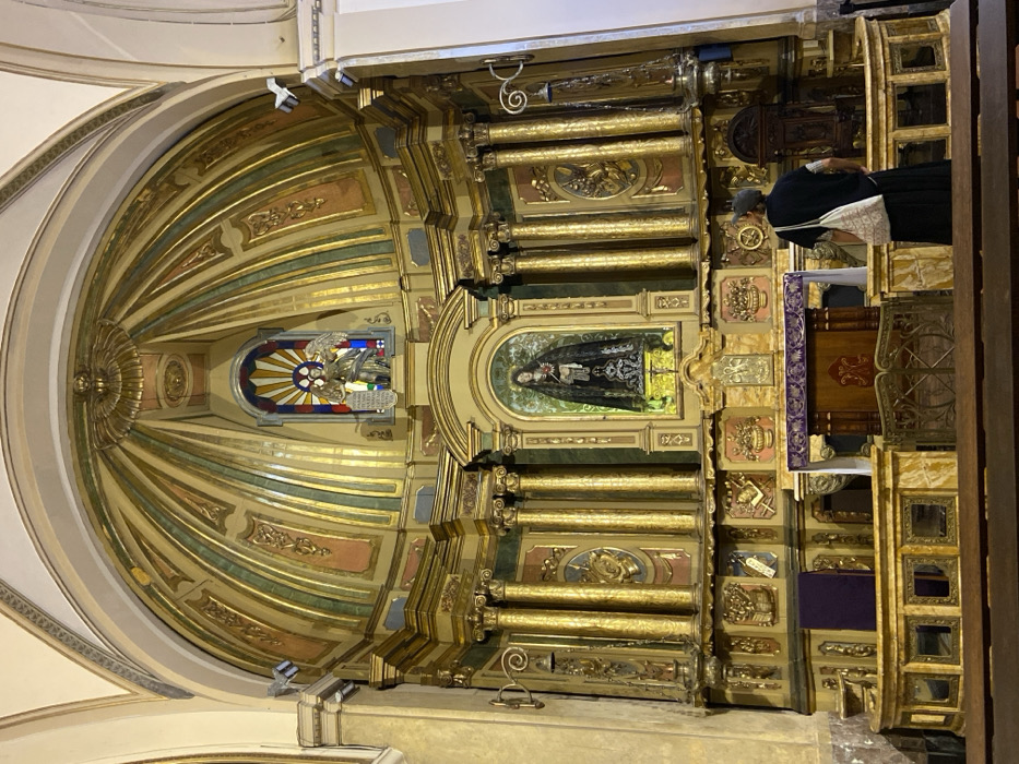 The image depicts the interior of the Basilica Nuestra Señora de la Piedad, located at Bartolomé Mitre 1524, Monserrat, Buenos Aires, Argentina. The central focus is a large, highly ornate, gilded altarpiece dominating the scene.
The altarpiece features multiple tiers of architectural elements, primarily rendered in gold leaf, with fluted columns, intricate carvings, and decorative panels in muted greens and pinks. At its center, within a glowing niche, stands a statue of Our Lady of Sorrows (Mater Dolorosa), clad in a dark, draped garment, with a red heart pierced by swords visible on her chest. Above this, within an upper arched recess framed by the gilded structure, a statue of an winged angel (likely St. Michael) holding a shield and sword is positioned in front of a semicircular stained-glass window depicting a cross and a sunburst pattern.
In front of the altarpiece, an altar table is draped with a purple liturgical fabric, partially obscured by a low railing constructed of marble and decorative metalwork. To the lower right, an adult individual is visible from behind, standing and facing the altarpiece. This person wears a dark long-sleeved top, a dark long skirt or trousers, and a dark cap or head covering. A light-colored, patterned tote bag hangs over their right shoulder. In the immediate foreground, the tops of dark wooden pews are visible, suggesting the perspective is from the nave of the church. The upper walls and ceiling beyond the altarpiece are light-colored with subtle architectural moldings, and artificial light fixtures are mounted on the upper side walls.