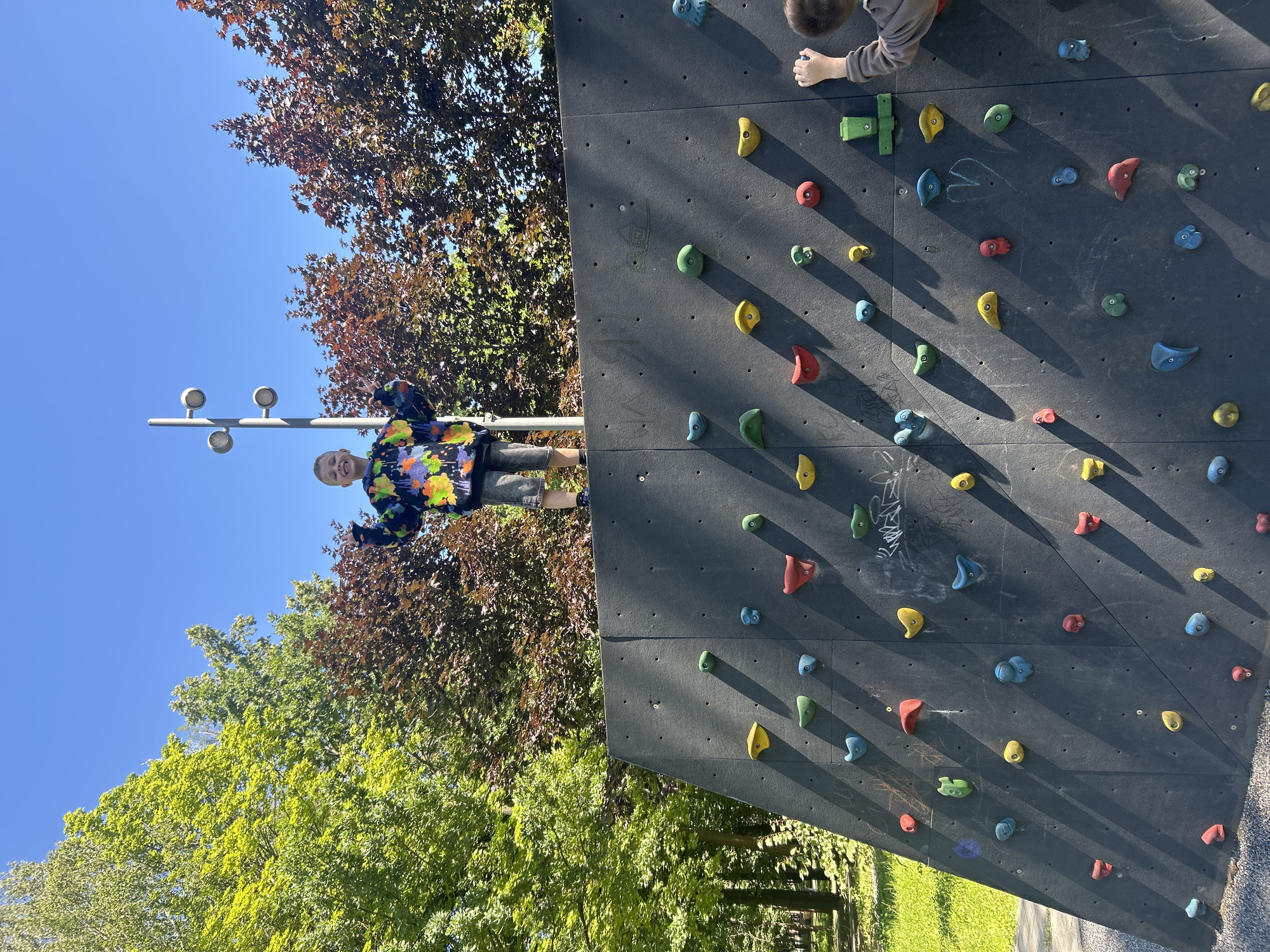In Khimki, Russia, a young boy, possibly around 7-10 years old, stands triumphantly at the top of a climbing wall. He is wearing a colorful, tie-dye hooded sweatshirt and grey shorts. His arms are raised in a celebratory gesture, and he is smiling broadly. The climbing wall is black with numerous brightly colored holds of various shapes and sizes, arranged in a challenging pattern. Sunlight casts distinct shadows from the holds onto the wall, indicating a bright, sunny day. Behind the climbing wall, lush green trees and a clear blue sky create a vibrant outdoor setting. To the right of the frame, another child is partially visible, reaching for a hold on the climbing wall. The scene appears to be in a park or recreational area, with grass and more trees visible in the background.