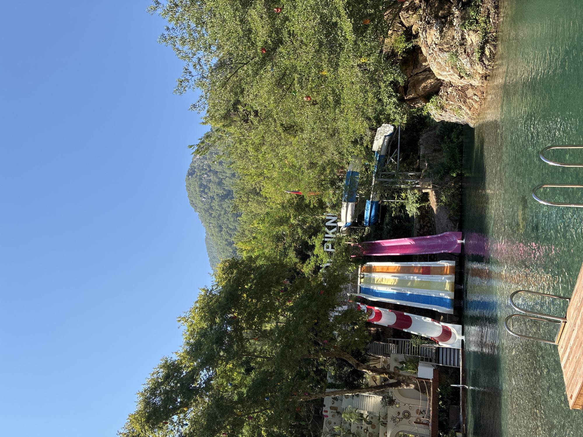 The image captures a natural water park setting nestled in a lush, mountainous landscape. In the foreground, a clear, turquoise body of water with a wooden deck and stainless steel ladder invites swimmers. Several colorful water slides, including red and white, blue and white, yellow and white, and pink, cascade down from a raised platform, their water streams visibly entering the pool. Behind the slides, dense green trees and foliage climb up the slopes of a rocky mountain under a clear blue sky. A white sign with dark lettering is partially visible among the trees, with the word "PIKN" decipherable. The overall impression is one of a vibrant, recreational outdoor space integrated with the natural environment. This location is identified as the Alanya Piknik area in Alanya, Turkey.
