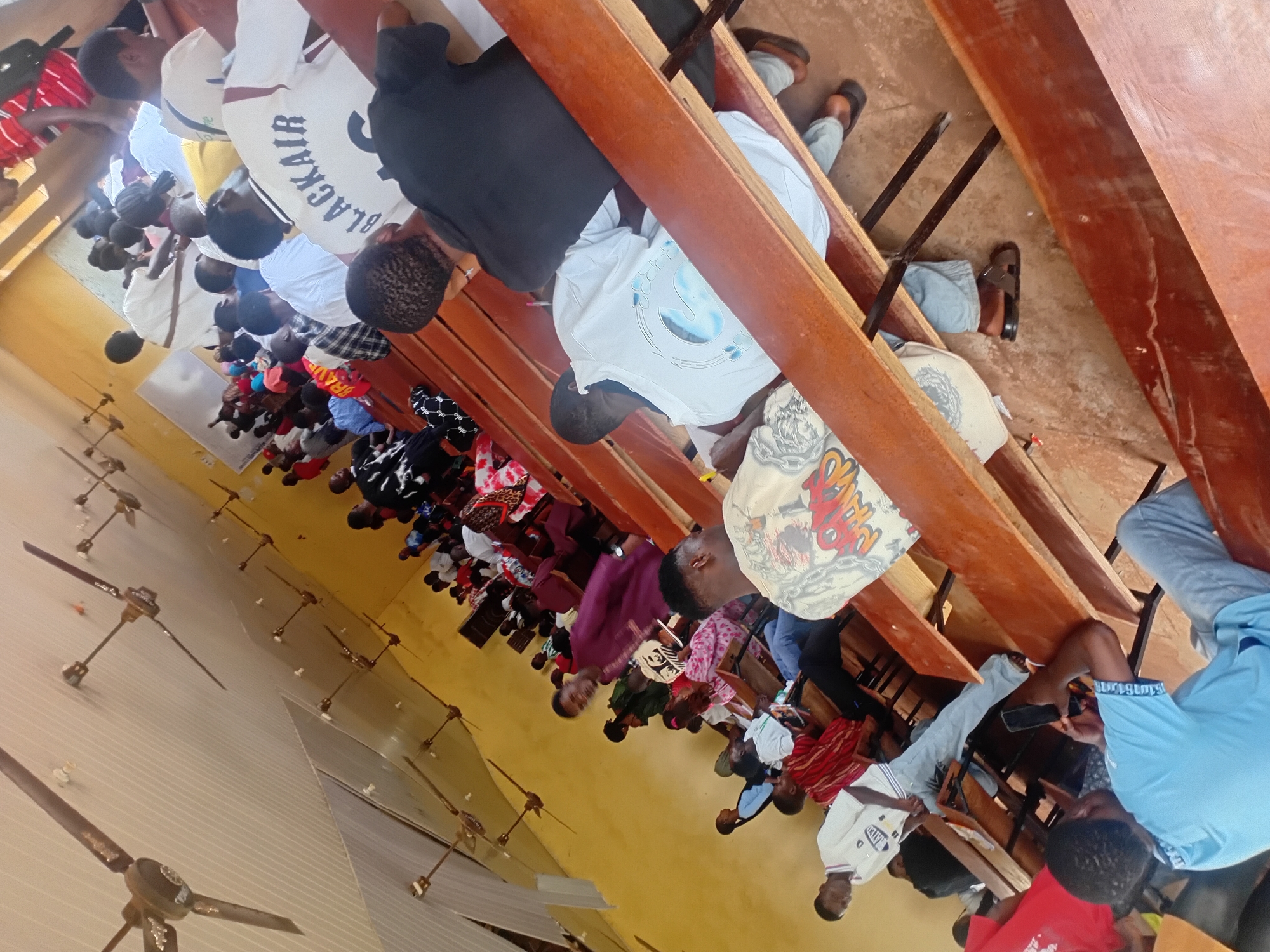 The image depicts a crowded indoor scene, likely a classroom or assembly hall in Ogbogbo, Nigeria. The room has a yellow-painted wall and a white ceiling with multiple ceiling fans. Long wooden benches arranged in rows serve as seating. Numerous individuals, predominantly young adults and teenagers, are present. Some are seated on the benches, while others appear to be standing or in motion.
In the foreground, a person wearing a light blue t-shirt is seated and looking at a mobile phone. To their right, another person in a white t-shirt with graphics on the back is seated, facing away from the camera. Further back, a person in a dark purple garment is standing and looking towards the front of the room. Many individuals are wearing casual clothing, including t-shirts, shirts, and pants. Some are dressed in more formal attire, like a person in a white shirt and a red and white striped shirt.
The atmosphere appears to be one of an educational or religious gathering. The presence of numerous people and the setting suggests a community event or a lesson. The lighting is a mix of natural light coming from an unseen source and the light from the ceiling. The floor is visible in some areas and appears to be a packed dirt or concrete surface. The overall scene conveys a sense of activity and congregation.