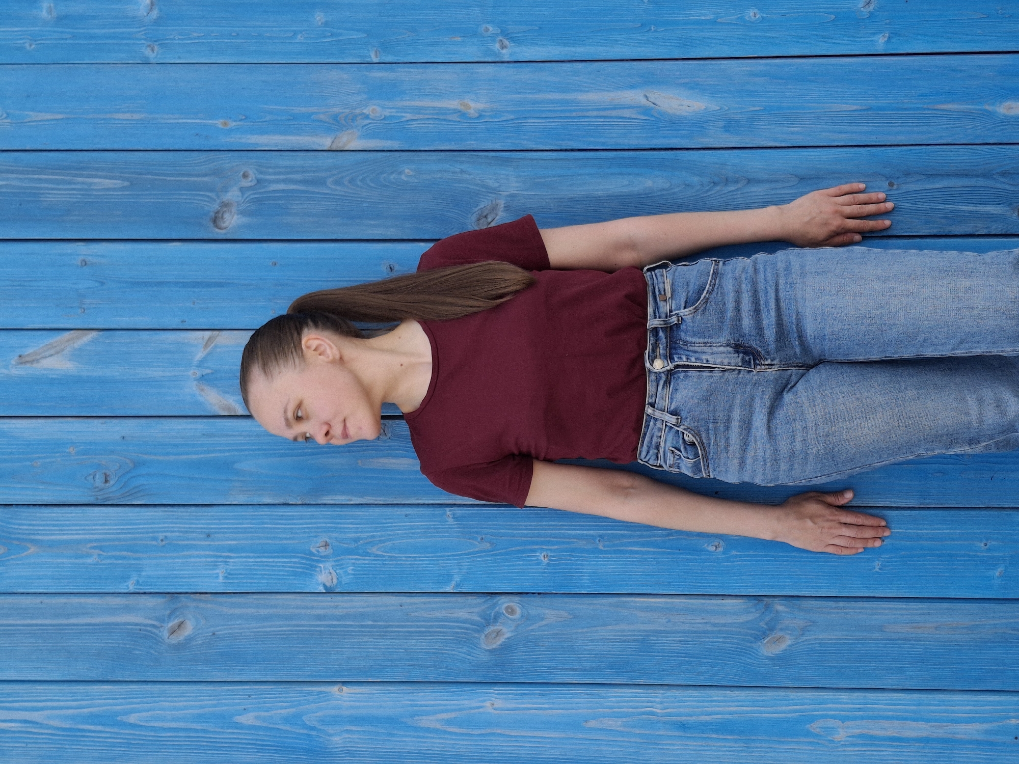 A young woman is lying on a wooden surface painted blue, which appears to be an exterior wall. She is wearing a maroon t-shirt and light blue jeans. Her hair is pulled back in a ponytail. The woman's body is oriented horizontally across the frame, with her head on the left and feet on the right. The location is Chaikovsky, Russia.