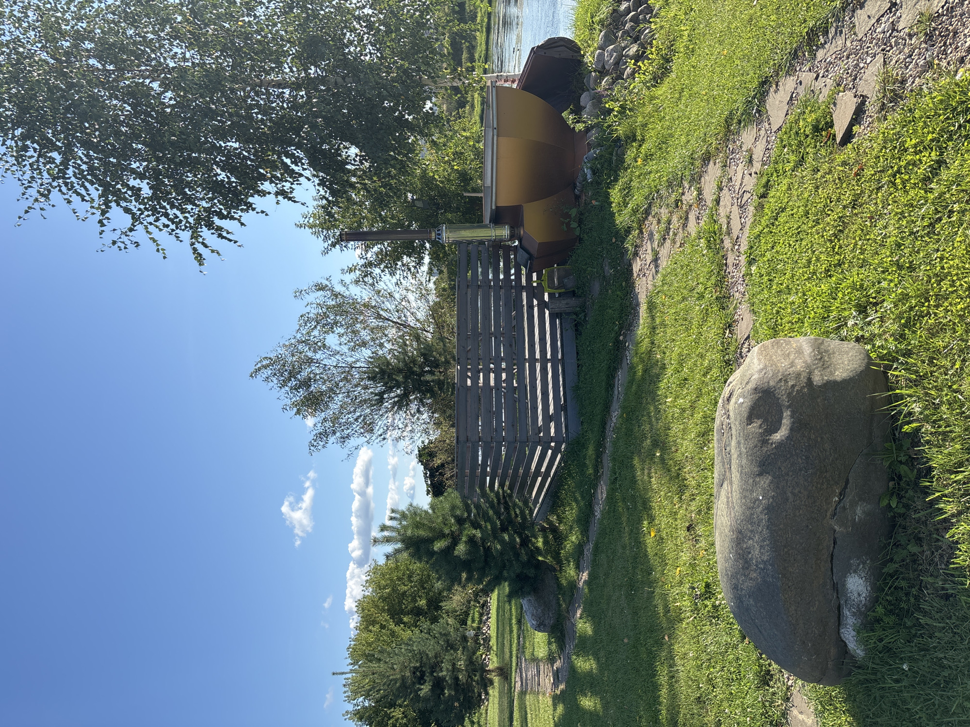The image depicts an outdoor scene on a sunny day with a clear blue sky and scattered white clouds. In the foreground, a large, weathered boulder rests on a patch of green grass, intersected by a stone pathway. Behind the boulder, a wooden deck with a grey slatted fence surrounds a large, metallic, cauldron-like structure with a chimney. This structure appears to be a hot tub or a wood-fired bath. To the left of the deck, two evergreen trees stand tall, with a third larger deciduous tree to the right, its branches extending towards the sky. In the background, a calm body of water, possibly a river, flows gently, reflecting the sunlight. The overall environment suggests a tranquil, natural setting, likely a backyard or a recreational area, possibly located in the vicinity of Zvenigorod, Russia, given the architectural style of the outdoor elements and the presence of a water body and vegetation. There are no people visible in the image.