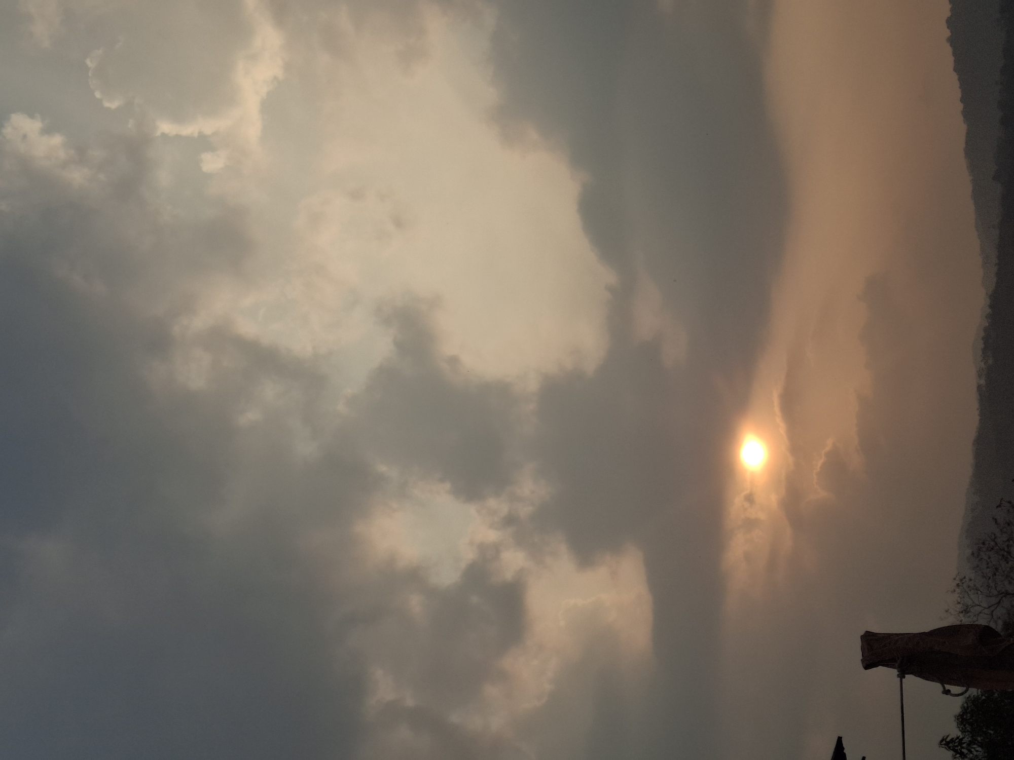 An overcast sky with the sun faintly visible behind the clouds, casting a soft, warm glow. The sun is positioned towards the lower left quadrant of the image, appearing as a bright, hazy orb. The clouds are a mix of dark grays and lighter, diffused tones, suggesting either early morning or late afternoon light. In the foreground, the silhouette of a rugged, possibly artificial structure is visible on the left, with a thin horizontal bar extending from it. Bare branches of trees are also discernible in the lower portion of the frame. The background features a dark, undulating line of hills or mountains, softened by the atmospheric conditions. The overall impression is a serene, natural landscape with a hint of human presence.