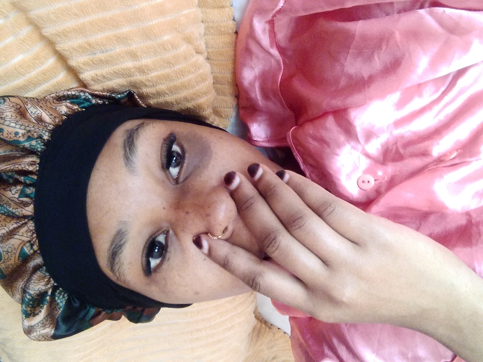 The image depicts a close-up, rotated view of a person lying down. The person is wearing a pink silk garment, possibly pajamas or a robe, which is partly visible on the left side of the frame. They are also wearing a black bonnet with a patterned trim. The person's face is partially obscured by their hand, which is covering their nose and mouth. A subtle nose ring is visible. The background consists of a textured, light orange surface, likely a blanket or bedding. The lighting suggests an indoor setting. The image does not contain any information to determine its location in Nyeri, Kenya, or any other specific geographical area.