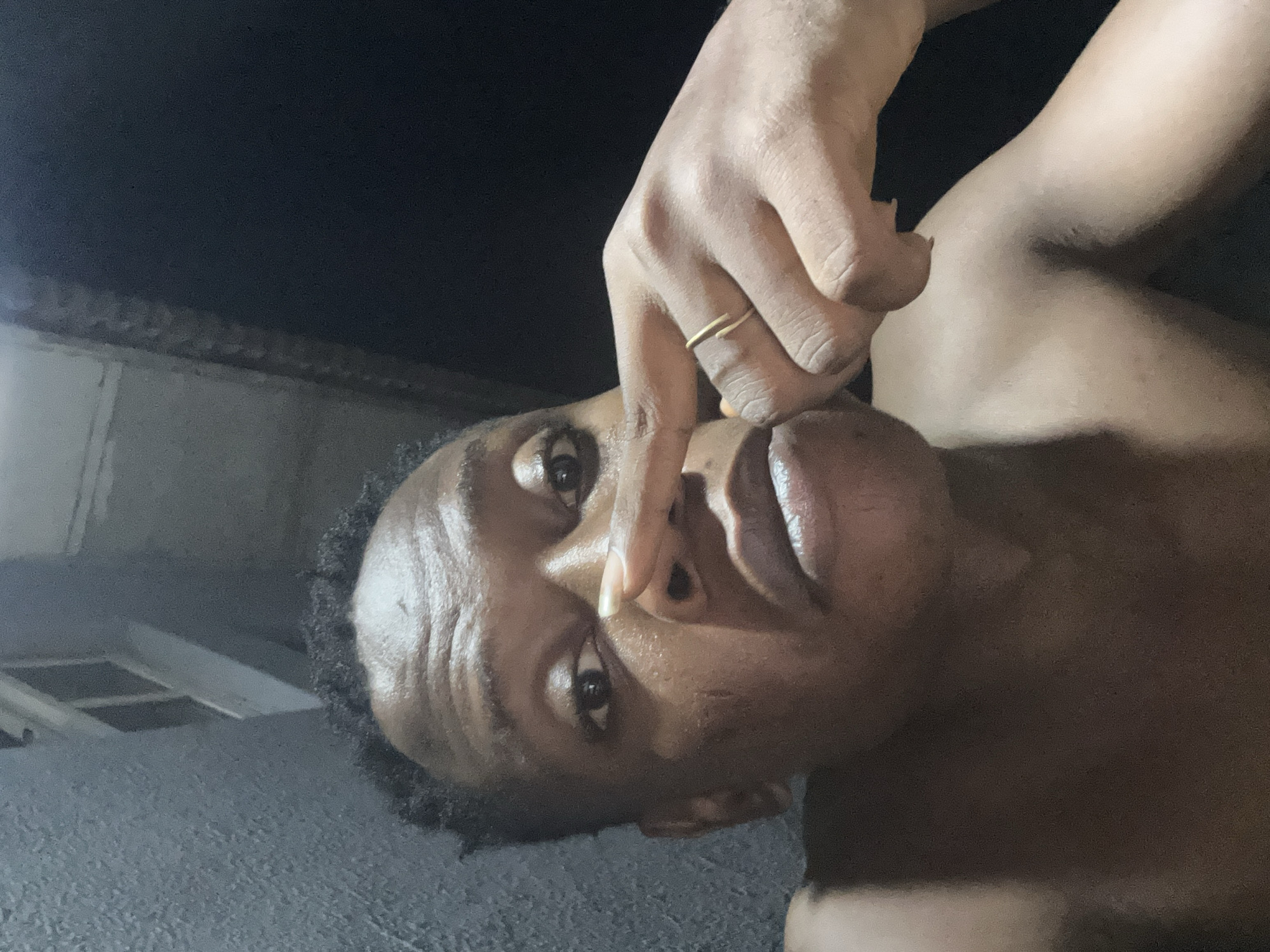 This image, identified as originating from Ikorodu, Nigeria, features an adult individual centered in a vertical composition. The person, appearing male, has a dark skin tone, short dark hair, and is depicted from the upper torso upwards. The individual is shirtless. Their left index finger is pressed against the bridge of their nose, with the palm of the left hand visible. A simple, thin gold-colored ring is worn on the ring finger of the left hand. The individual's eyes are open and directed towards the camera, with a neutral to slightly pensive facial expression. A faint scar or mark is visible on the left cheek, below the eye.
The background is split. To the viewer's left, a light grey, textured wall, possibly concrete or stucco, is visible. Above the individual's head, on the left side of the frame, an architectural detail appears, showing a light-colored structure with what resembles a window frame. The right side of the background is completely dark, indicating a lack of illumination or a deeply shadowed area. The subject is unevenly lit by a light source seemingly positioned to the upper left (from the viewer's perspective), creating highlights on the forehead and casting shadows beneath the nose and chin.