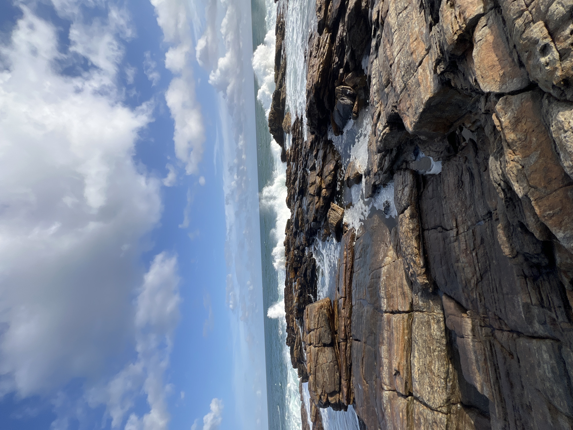 The image depicts a rocky coastline with waves crashing against the shore. The rocks in the foreground are wet and appear to be a reddish-brown color, with some pools of water reflecting the sky. In the background, the ocean is a deep blue, with white foam from the breaking waves. The sky is a bright blue with scattered white clouds. The scene suggests a natural, undeveloped coastal area. The location is identified as Kadurupokuna, Sri Lanka. There are no people or discernible man-made objects in the image.