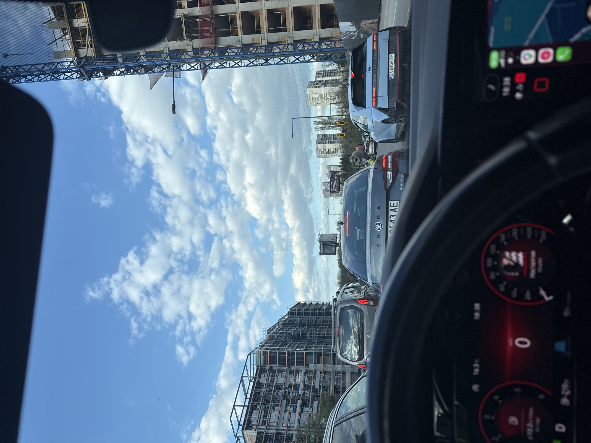 The image, taken from inside a vehicle, captures a street scene in Sofia, Bulgaria, during what appears to be a traffic jam or stopped traffic. The perspective is from the driver's seat, with parts of the steering wheel and dashboard visible in the foreground on the right. The dashboard digital display shows the time as "18:38" and a speed of "0," indicating the vehicle is stationary. A mounted smartphone to the left of the dashboard also displays "18:38" and various app icons.
In front of the observer's car, two other vehicles are clearly visible. Immediately ahead is a dark gray Skoda Superb, with its rear and license plate "7643 AE" partially visible. In front of the Skoda is a light blue Hyundai Ioniq 5, displaying the Bulgarian license plate "EA 6905 AE". The "HYUNDAI" logo is visible on its rear. Further down the road, more cars are queued.
The background features a mix of urban development and residential areas. On the left, a prominent construction site with a large blue tower crane dominates the view, alongside several multi-story buildings under construction, characterized by concrete frames and extensive scaffolding. On the right, behind the parked cars, there are older multi-story residential buildings and more construction visible. The sky is bright blue with scattered white clouds, suggesting good weather. Other details include streetlights, a traffic light, and a small advertising board. The overall impression is that of an urban environment experiencing rush hour or a temporary halt in traffic.
