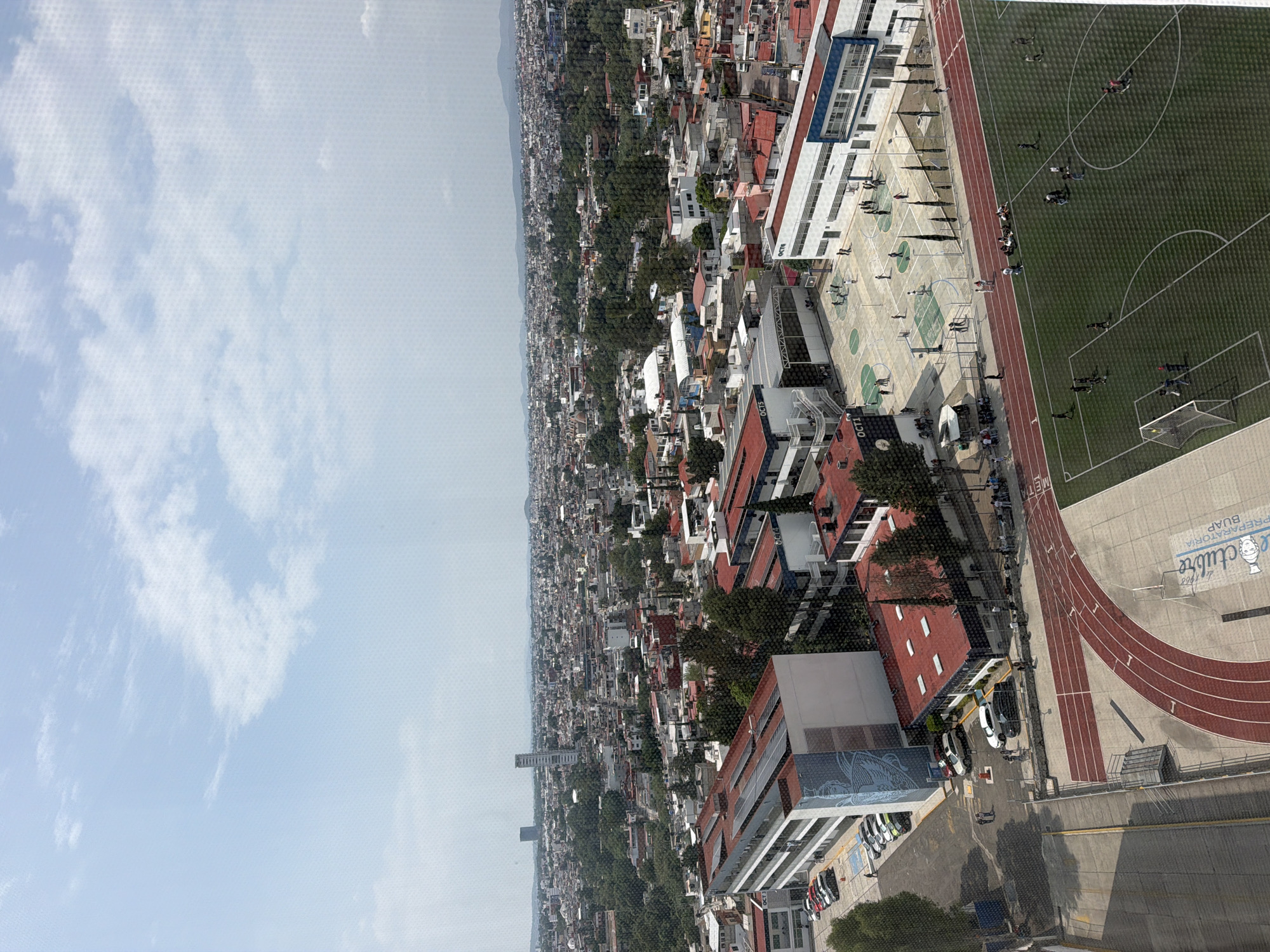 The image is an aerial view of San Bernardino Tlaxcalancingo, Mexico, featuring a school campus. The school is situated in a densely populated urban area characterized by residential buildings with red tile roofs interspersed with trees. The campus has a sports complex with a soccer field, a running track, and basketball courts. People are present on the soccer field and basketball courts. The sky is partly cloudy.