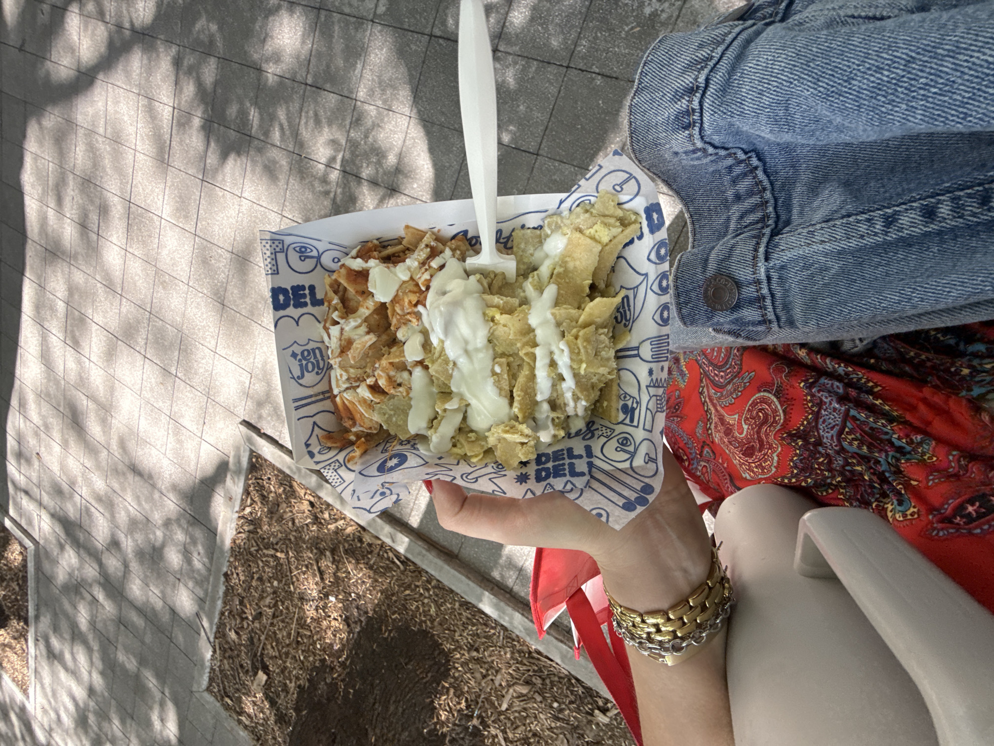 The image captures a first-person, high-angle view of a person holding a tray of food outdoors in Mexico City, Mexico. The subject's hand, adorned with gold and silver bracelets, cradles a white paper tray filled with what appears to be chilaquiles. The dish is segmented into two types, both covered in a generous drizzle of white cream or sauce. One side features smaller, reddish-brown pieces, possibly shredded chicken or crispy tortilla, while the other contains larger, softer tortilla pieces. A white plastic fork or spork is centrally placed in the food.
The paper tray is decorated with blue illustrations and text, including "DEL", "en Joy", "Tee", a stylized eye, and symbols of cutlery.
The person is wearing a denim jacket on their left arm and a vibrant red garment with a prominent paisley or floral pattern, likely a dress or skirt.
The setting is an outdoor, sunlit area. The ground consists of light-colored, square-tiled pavement, which is dappled with distinct shadows from trees or leaves, indicating a bright daytime. To the lower left, an area of dark mulch or soil is visible. The scene suggests a casual and enjoyable meal being consumed outdoors.