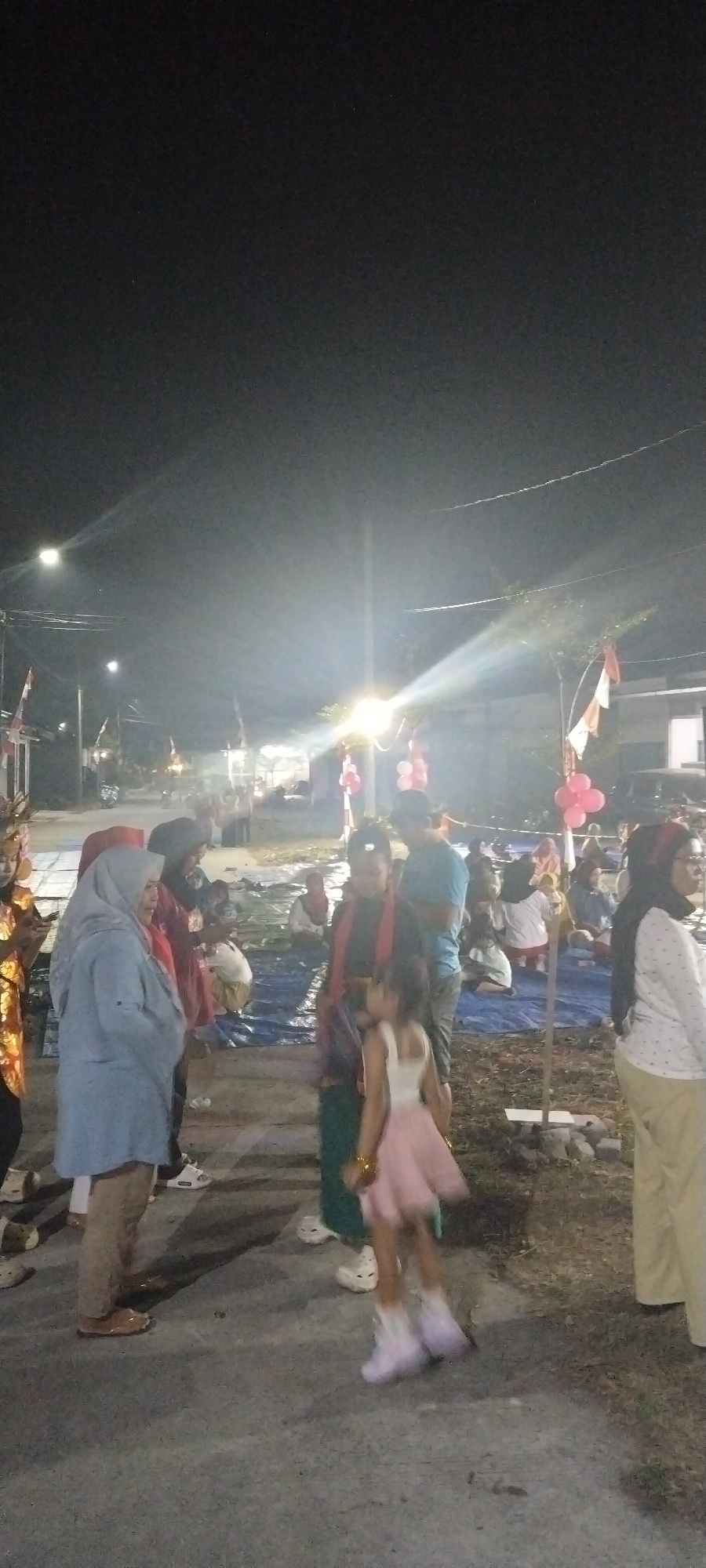 This image depicts an outdoor gathering at night, likely in Taban, Indonesia, given the presence of Indonesian flags. The scene is illuminated by artificial lights, creating a somewhat hazy atmosphere. In the foreground, a young girl in a pink dress and light-colored shoes is captured mid-motion, possibly dancing or playing. To her left, an adult in a light blue headscarf and beige pants stands, facing away from the camera. Several other individuals are visible in the mid-ground, some seated on blue tarpaulins spread on the ground, while others are standing and interacting. Background elements suggest a street or open area with buildings and trees, and some decorations like red and white balloons are present, hinting at a festive occasion. The overall impression is that of a community event taking place after dark.